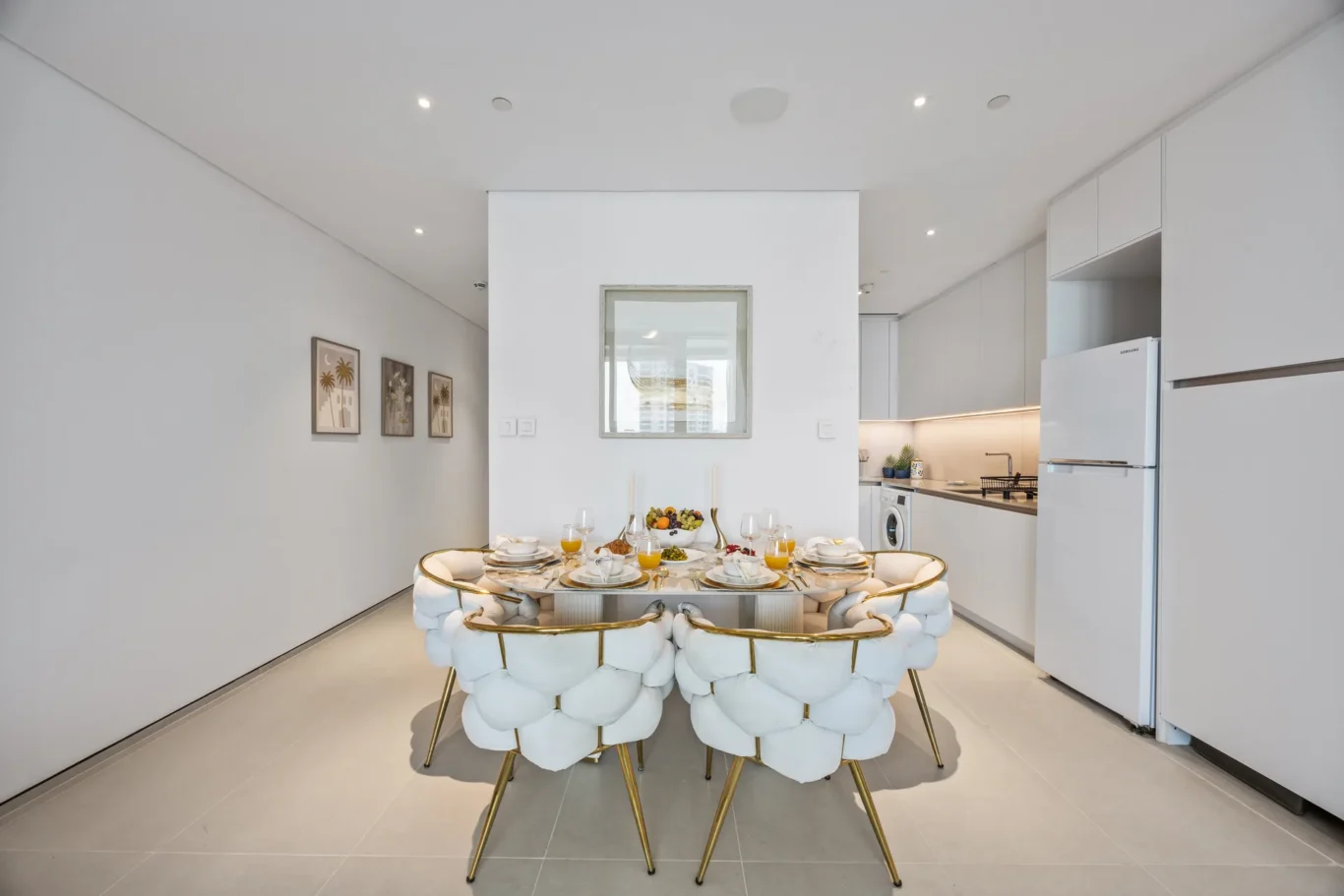 Modern white dining area with a round glass table and plush tufted chairs with gold legs; kitchen visible in background.