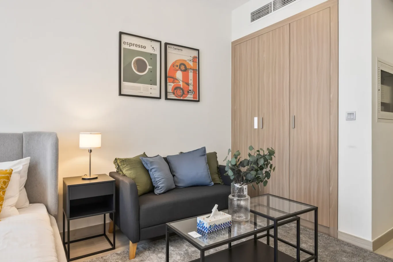 Modern living room with a gray sofa, decorative cushions, lamp, two framed wall posters, glass coffee table, and a tall wooden wardrobe.