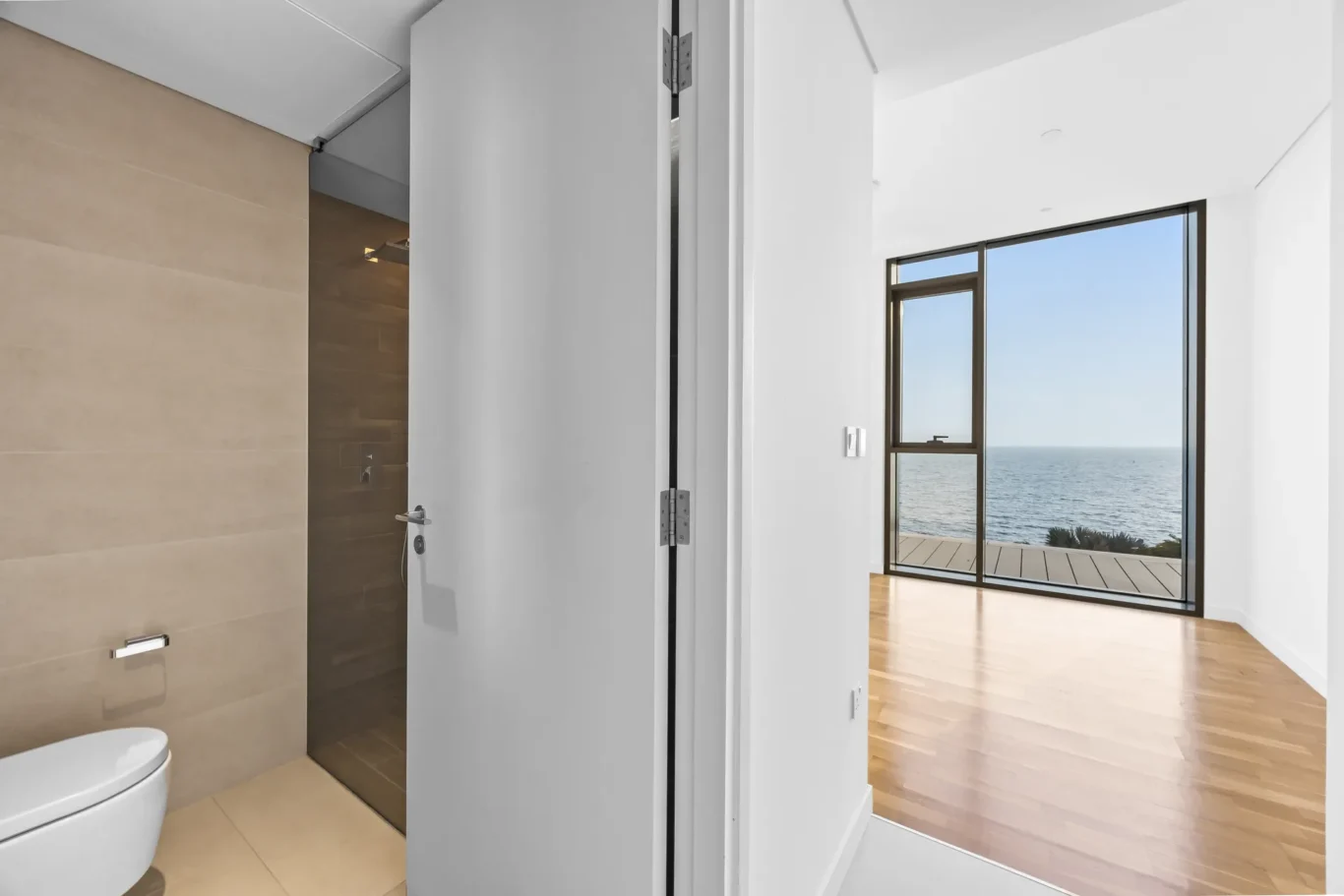 Modern bathroom with beige tiled walls and a visible toilet; an open doorway leads to a bright room with a large ocean-view window.