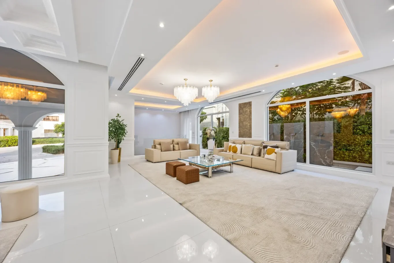 Luxurious white living room with beige sofas, glass coffee table, chandeliers, and large windows filling the space with natural light.