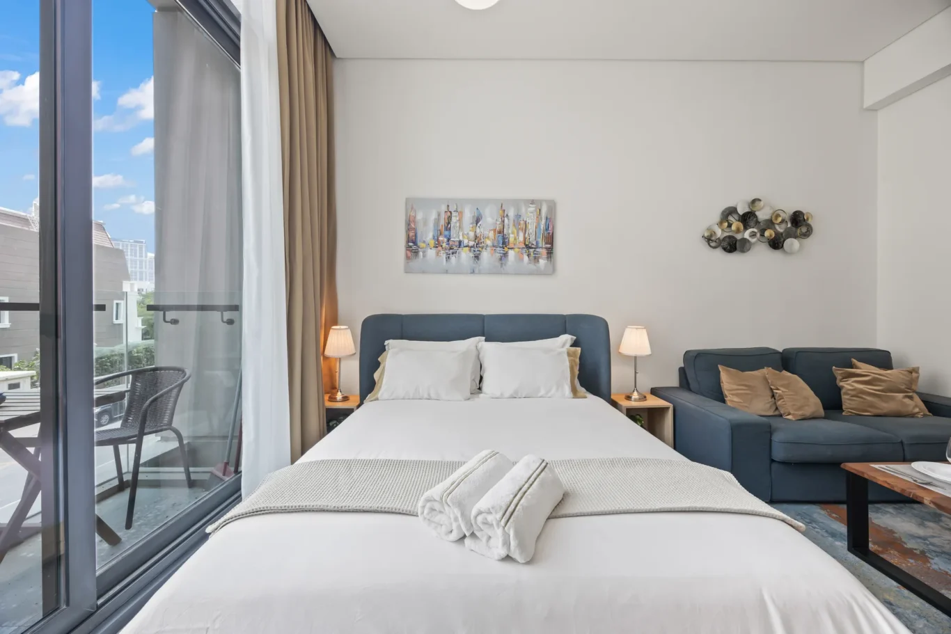 Hotel bedroom with a king bed, blue headboard, two nightstands, and a blue-gray sofa against a white wall; balcony doors open to a city view of buildings and blue sky.