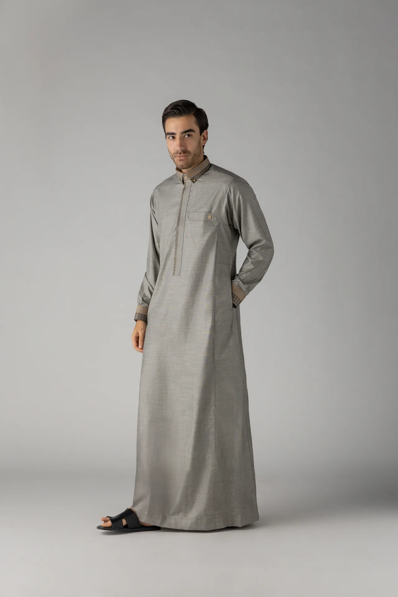 Man wearing a light gray traditional thobe and black sandals, standing against a gray studio backdrop, looking at the camera.],