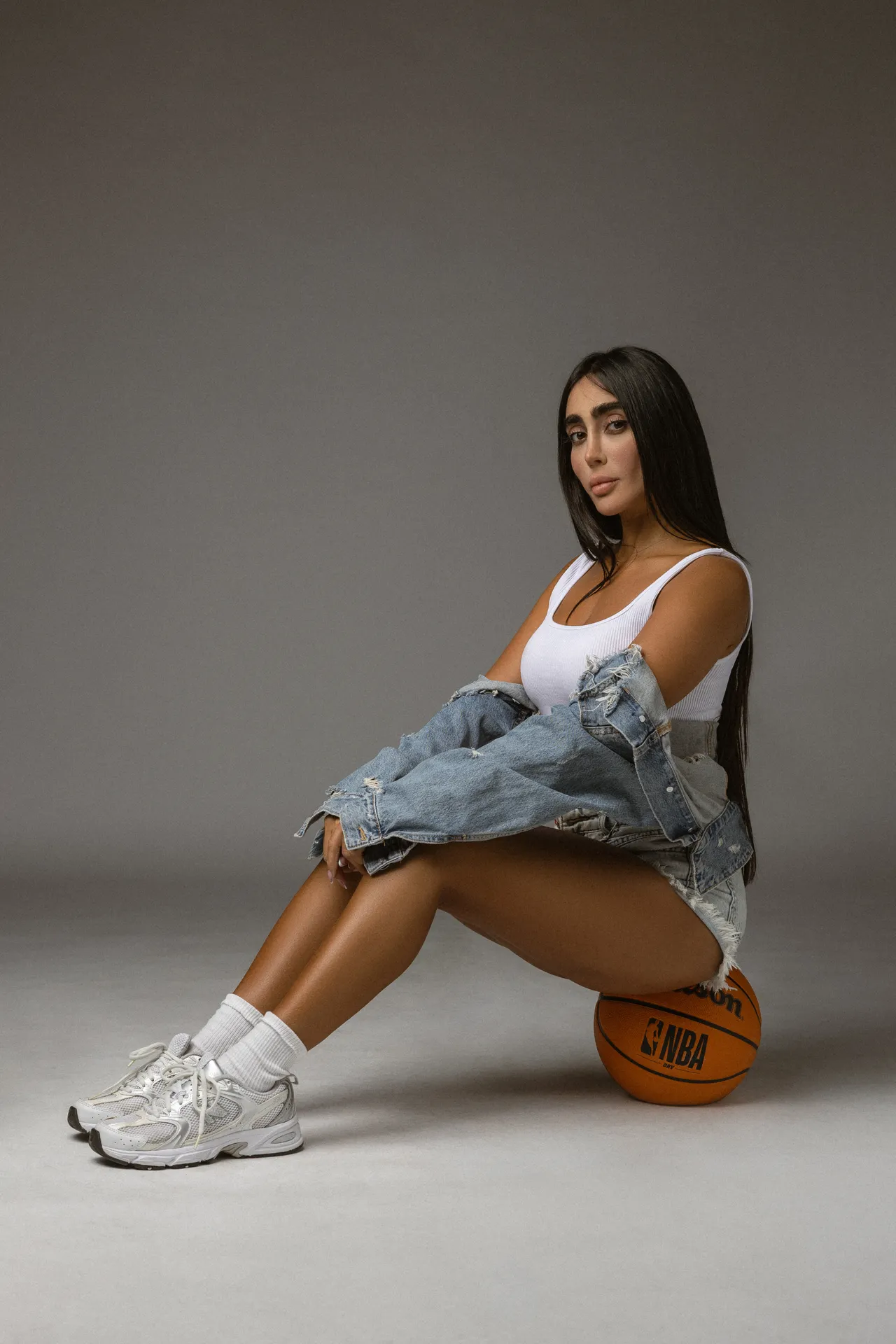 Woman seated on the floor with a basketball under her knee, wearing a white tank top, ripped denim jacket, and sneakers against a gray backdrop.