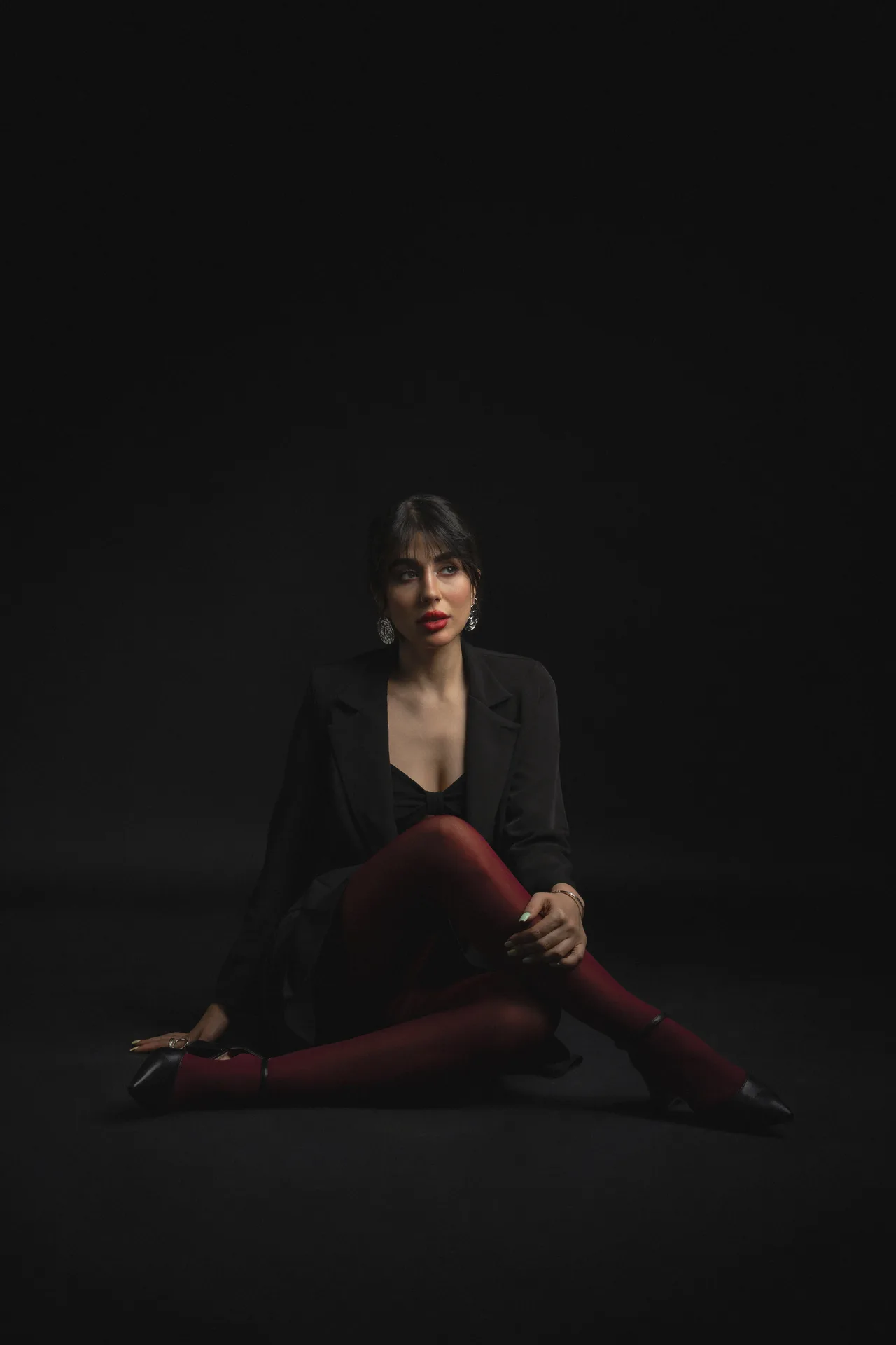 Fashion portrait of a woman in a black blazer and red tights seated on a dark studio floor, looking to the side with statement earrings.