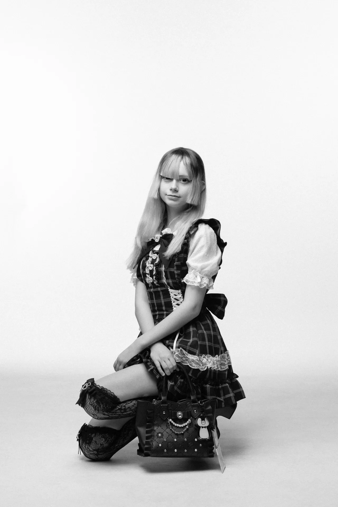Young person kneels on the floor in a plaid lace-trimmed dress, holding a decorative handbag in a studio setting (black and white).