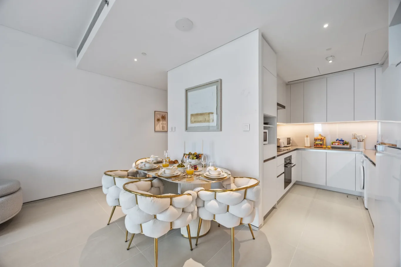 Luxurious white kitchen with a round, glass-topped dining table set for breakfast and tufted white chairs with gold legs on a light floor.