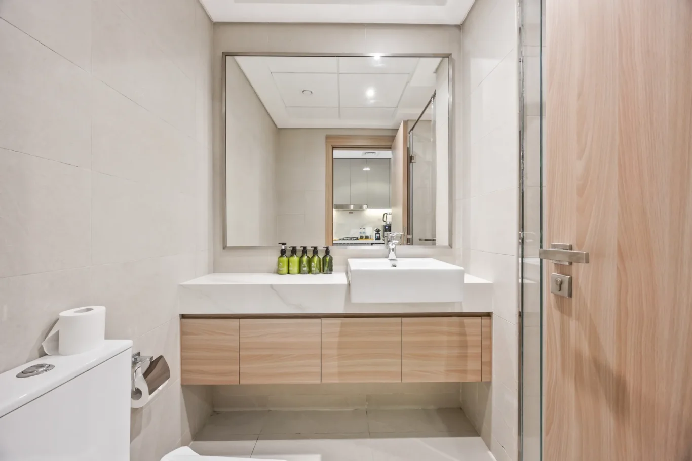 Modern bathroom with a white rectangular sink on a wood-toned vanity, a large wall mirror, and a row of green bottles on the counter.