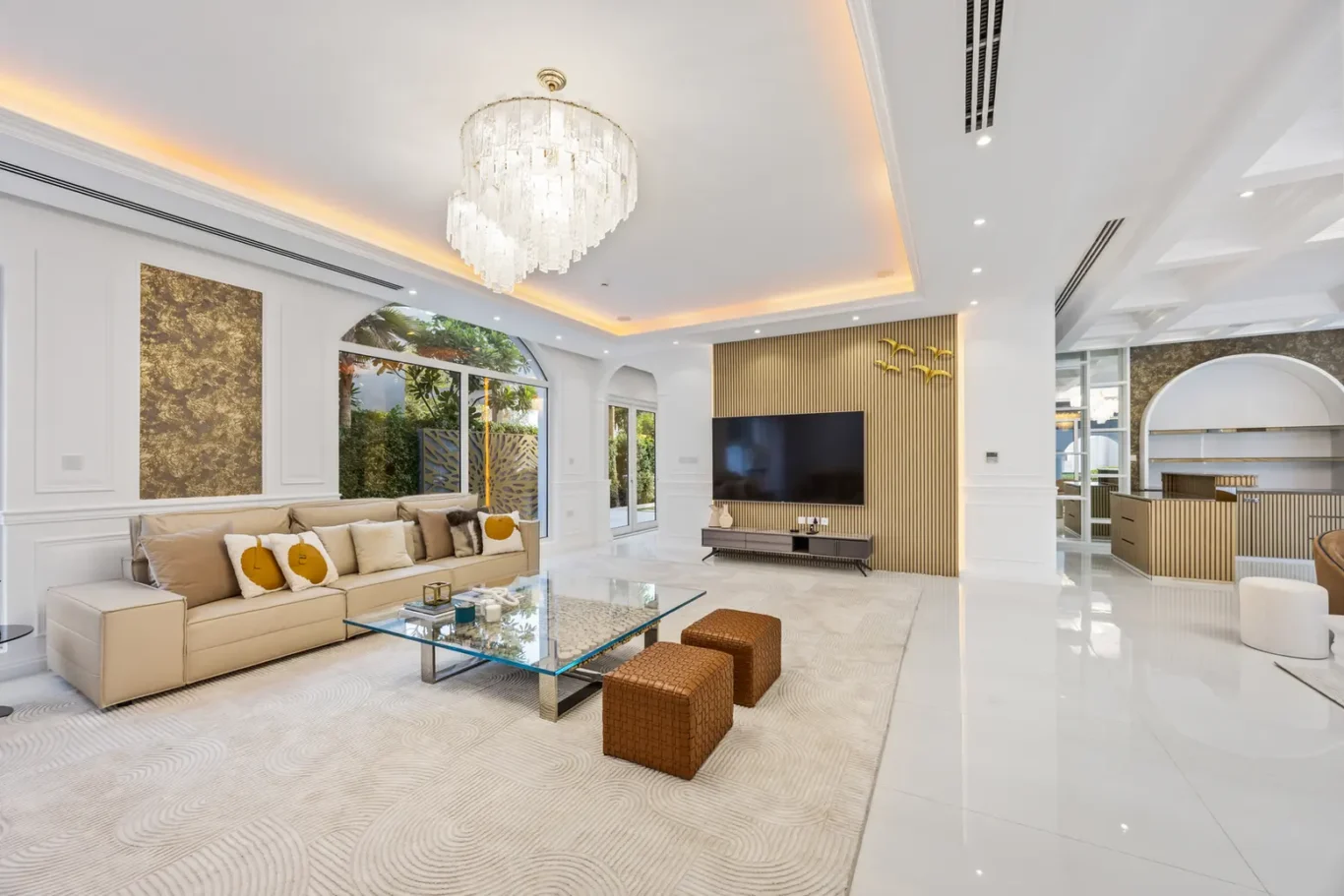 Luxurious bright living room with a large crystal chandelier, cream sofa, glass coffee table, and a wood-paneled TV wall.