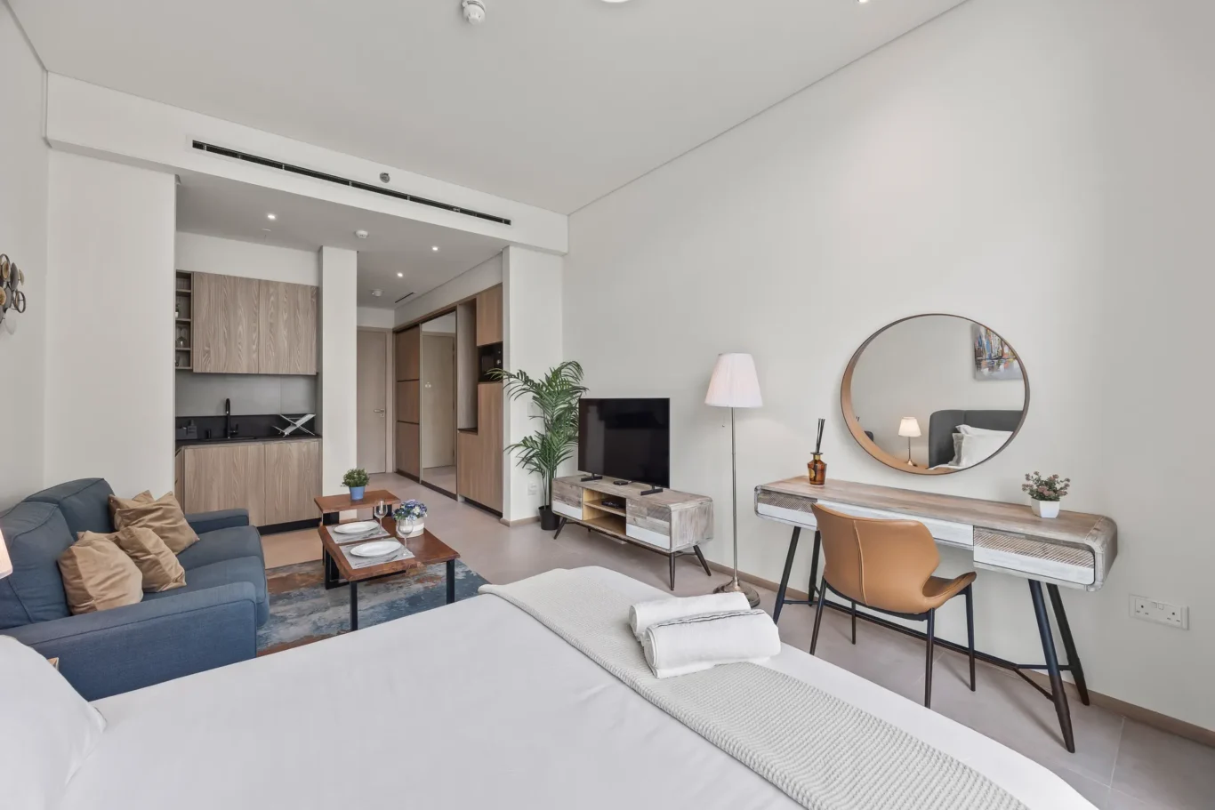Bright modern studio living area with a bed in the foreground, blue sofa, kitchenette to the left, and a round wall mirror.