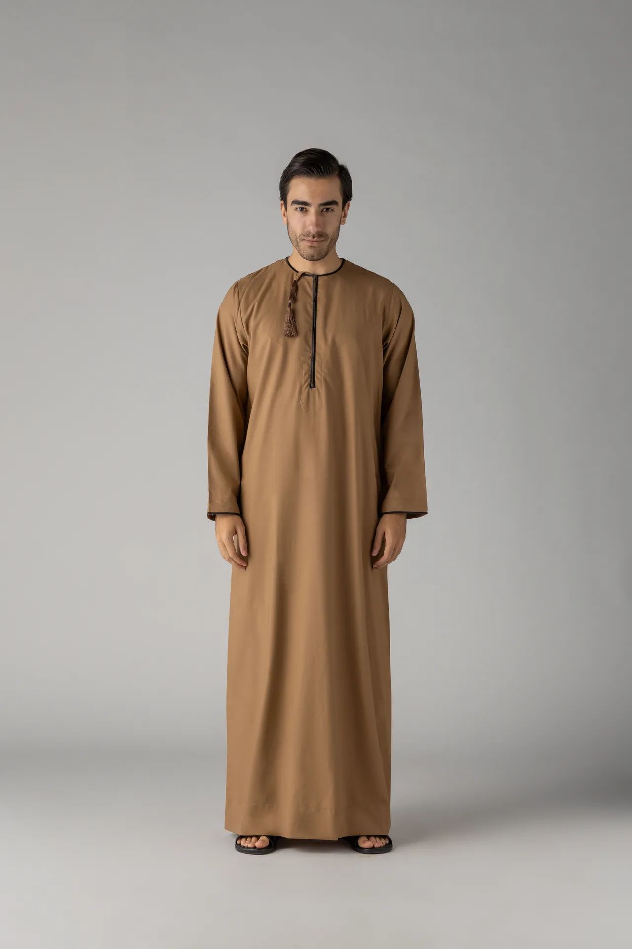 Front-facing portrait of a man in a long brown thobe with tassel, standing against a gray studio background and wearing sandals