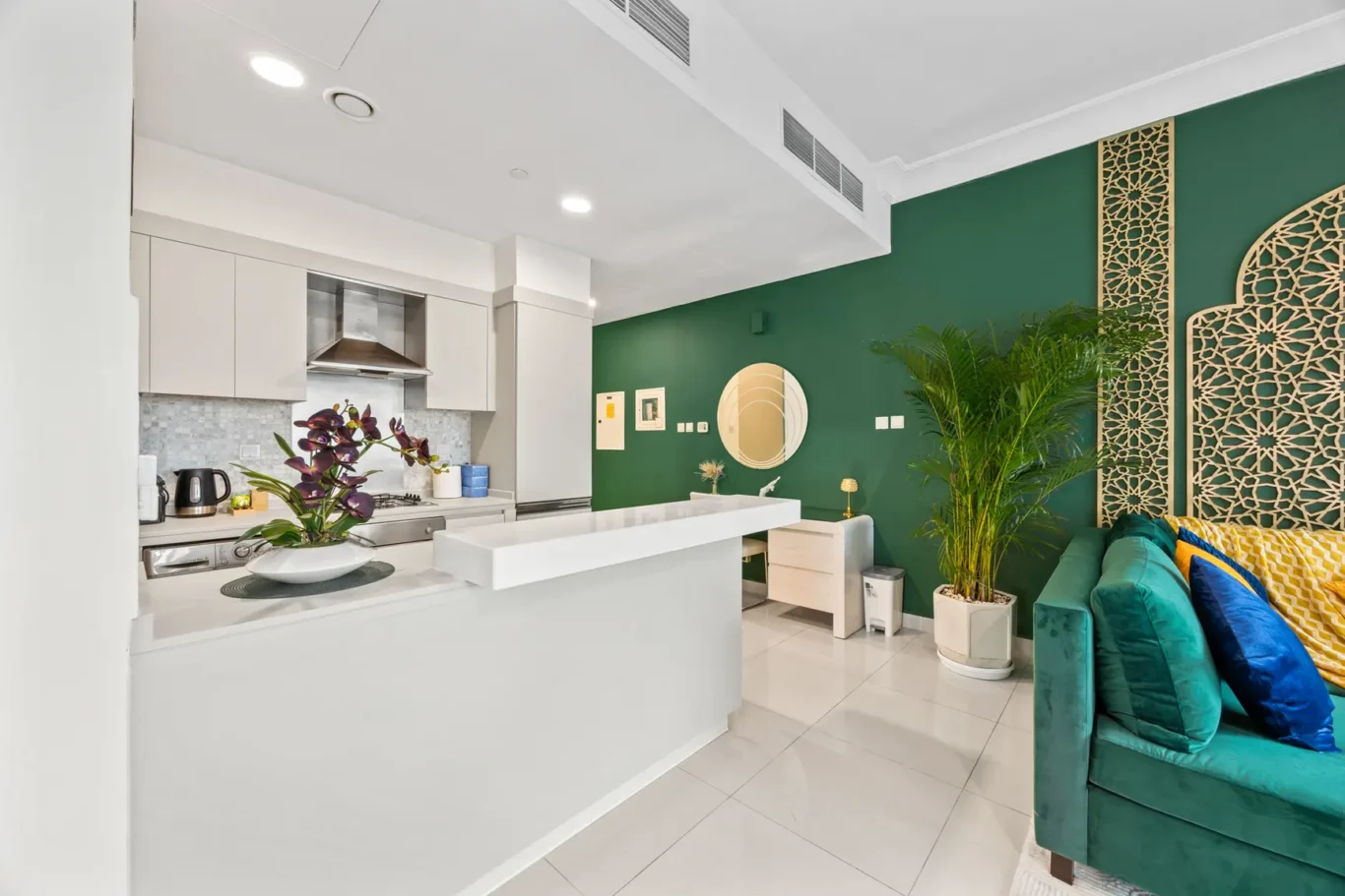 Open-plan living space with a white kitchen, green accent wall, decorative lattice panels, and teal sofa with colorful cushions.