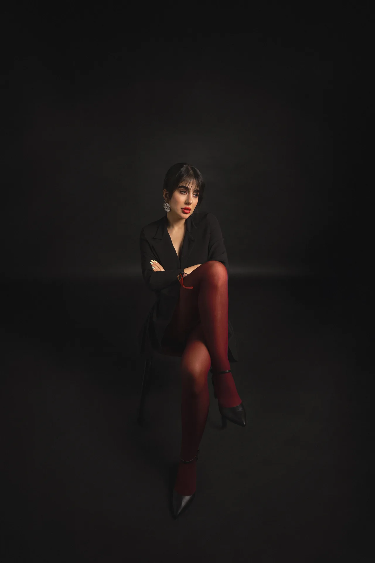 Portrait of a woman seated against a dark background, wearing a black blazer, red tights, black heels, bold red lipstick, and statement earrings.