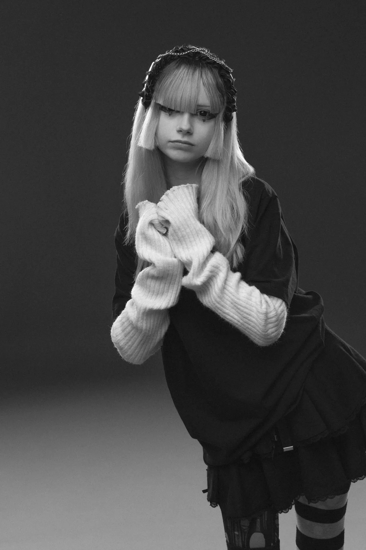 Black-and-white portrait of a person with long hair, wearing a lace headband and a chunky knit scarf wrapped around their arms, standing in a studio.