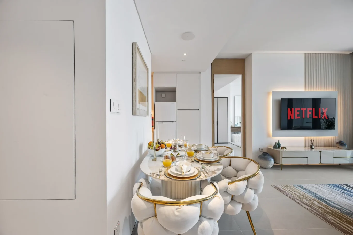 Open-plan dining and living area with a glass-topped round table set for breakfast, surrounded by plush white chairs with gold frames, and a wall-mounted Netflix screen in the living room.