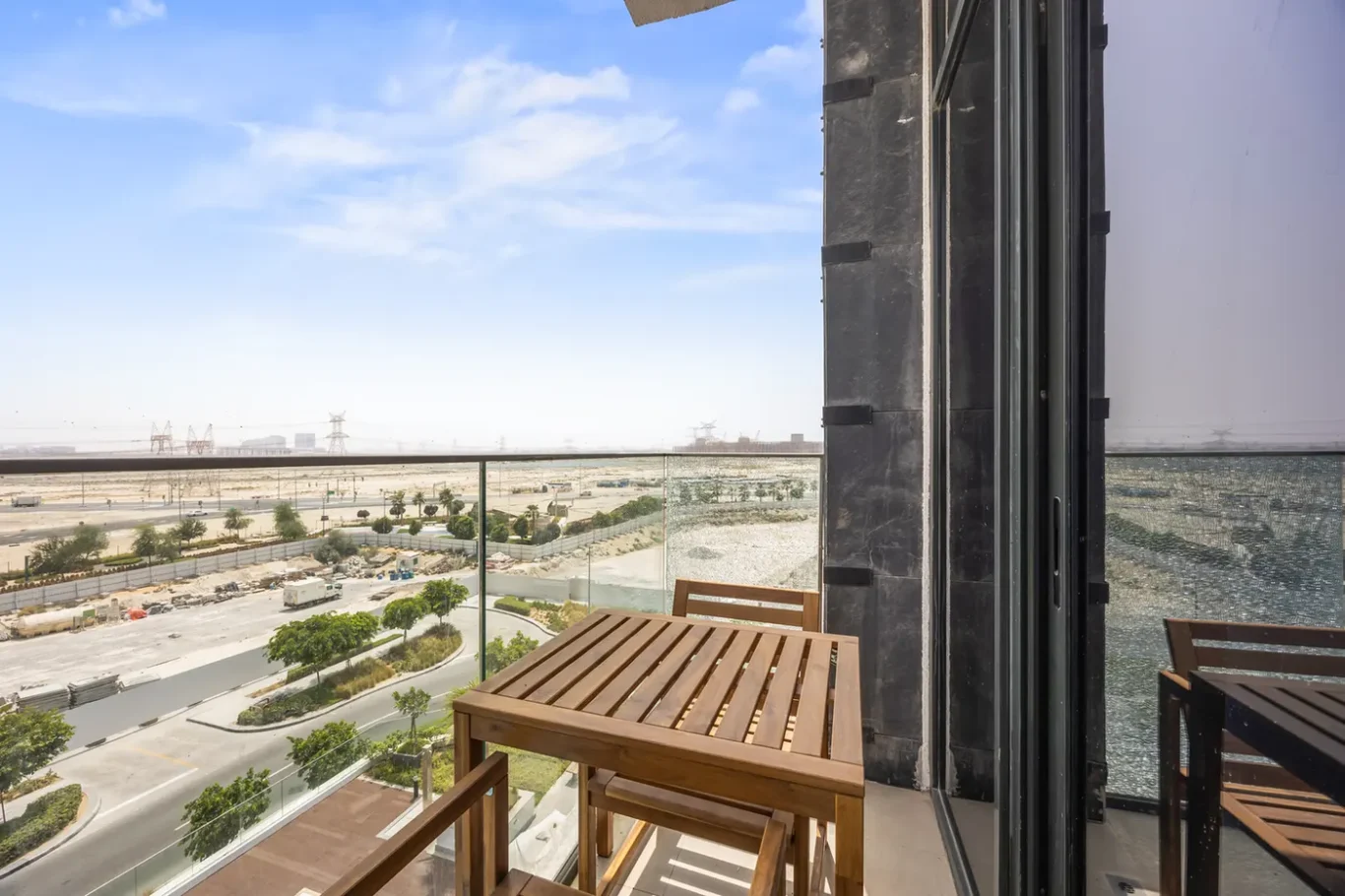 Wooden balcony table and chairs on a glass-railed balcony overlooking a desert road and distant power lines.