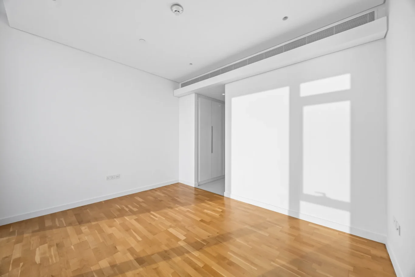 Bright empty white room with a light wood floor; a doorway to an elevator is visible in the back corner, with sunlight casting rectangular shadows on the wall