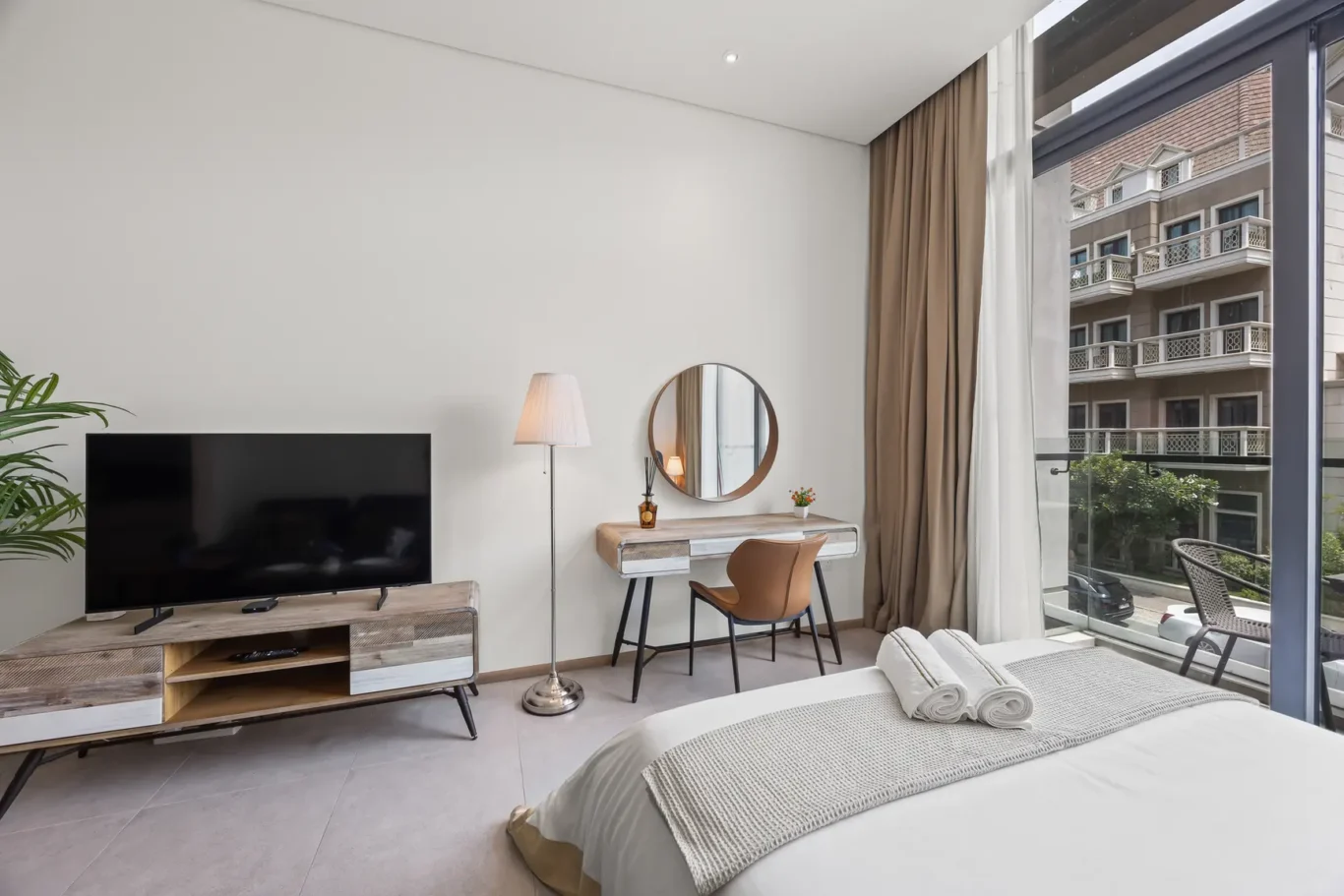 Bright hotel room with a king bed, wall mirror above a wooden desk, floor lamp, and a large window balcony view outside.