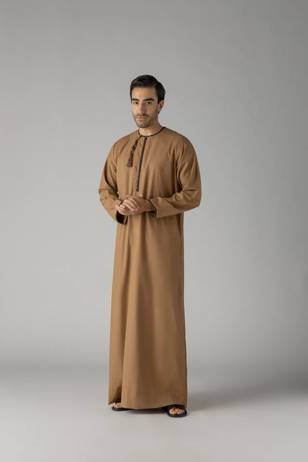 Man in a tan thobe with a tassel necklace standing with hands clasped in front against a gray background.