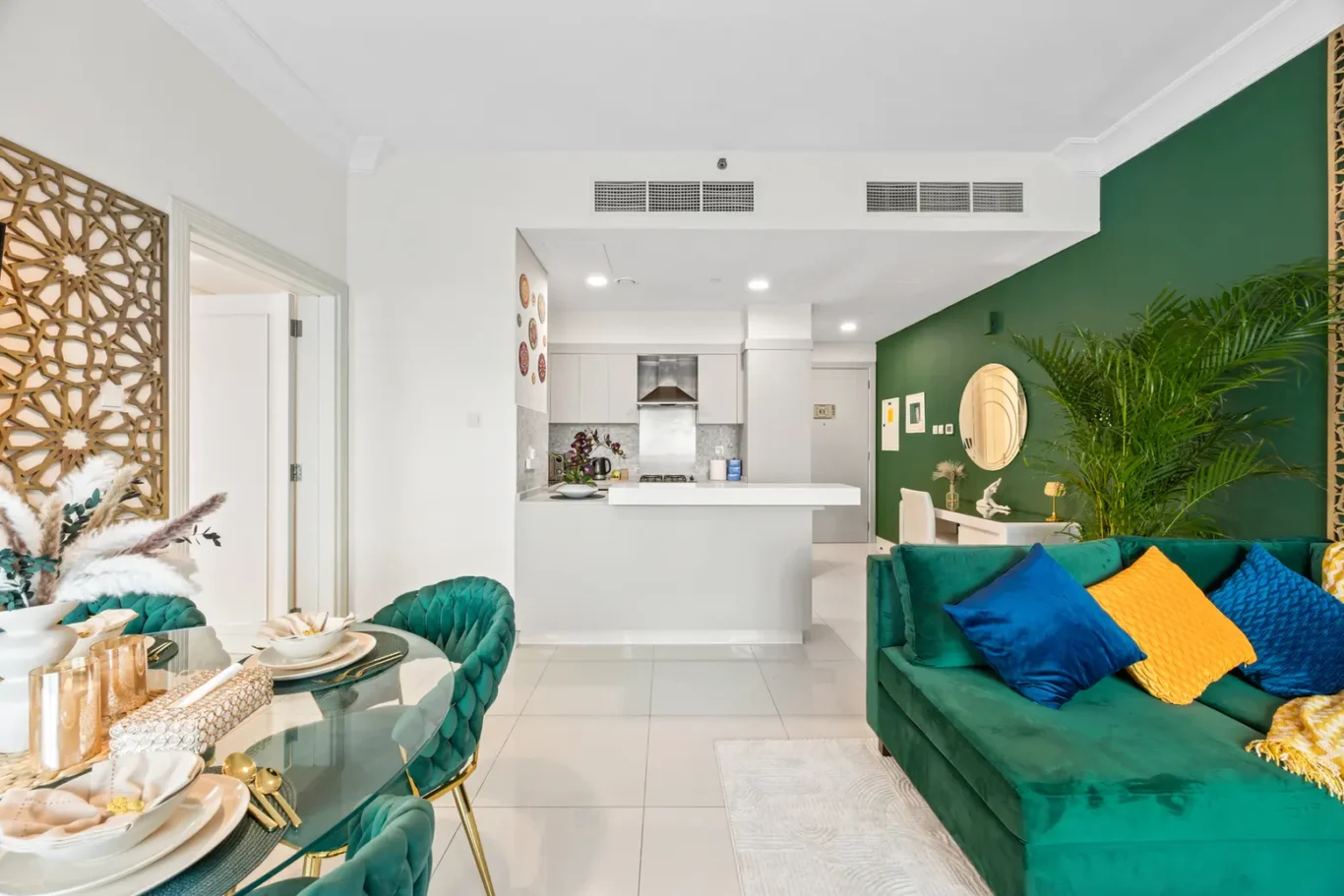 Open-concept living area: green accent wall, teal sofa with blue and yellow cushions, white kitchen in back.