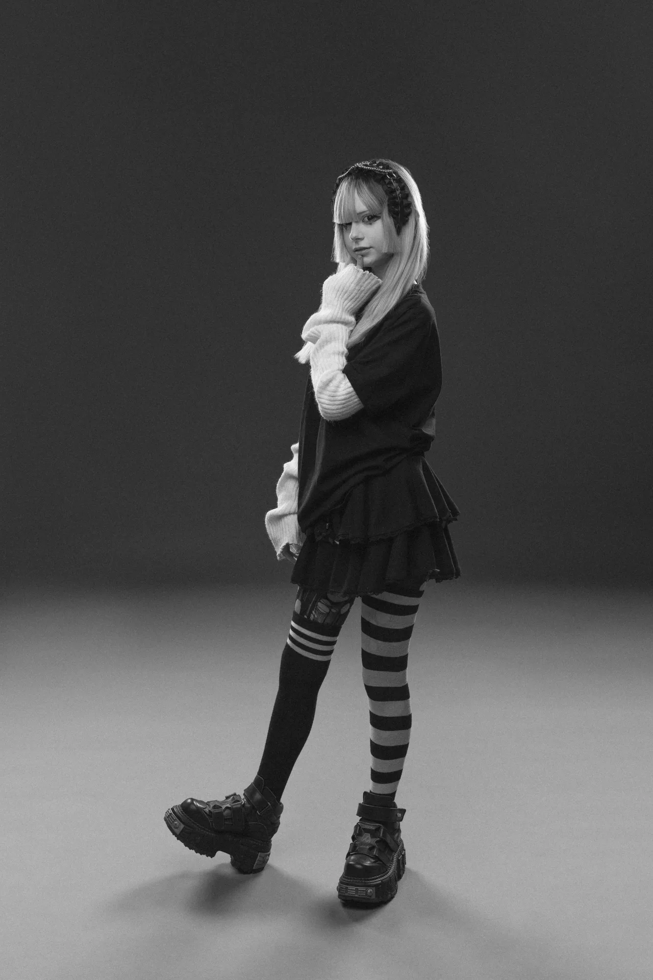 Stylish young woman in edgy black outfit with striped tights and chunky boots posing in a monochrome studio shot.