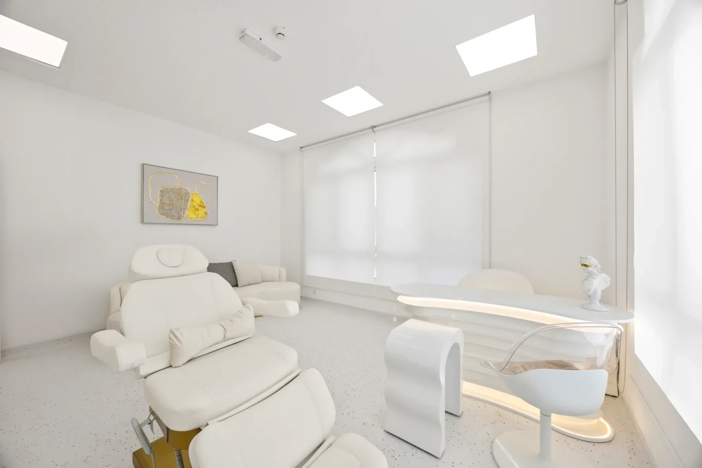 Bright, modern treatment room with white reclining chairs, abstract wall art, and a white curved table near a large window with blinds.