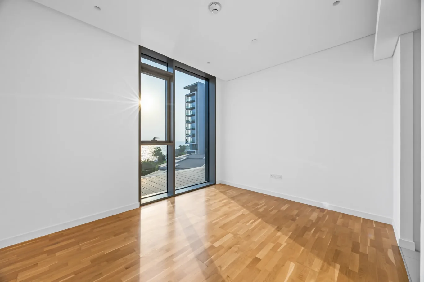 Bright empty room with large floor-to-ceiling glass doors opening to a balcony and an ocean view at sunset, wooden floor.