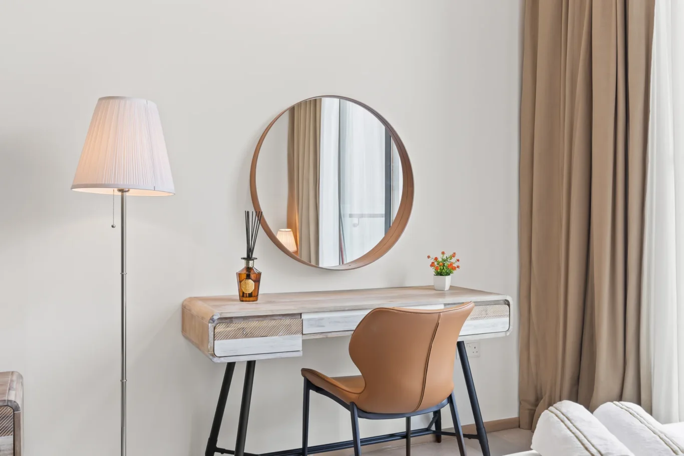 Round wooden-framed mirror above a softly lit vanity desk with drawers and a tan chair, in a calm bedroom.