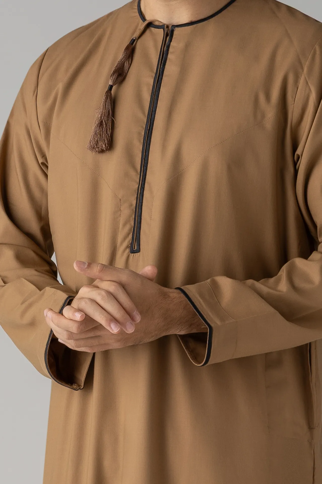 Close-up of a tan thobe with a black zipper and tassel at the collar, with hands lightly clasped in front.