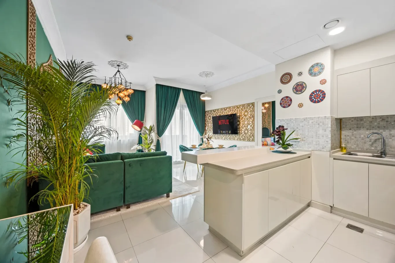 Open-plan living space with green sofa, tall plants, and teal curtains beside a white kitchen island and tiled backsplash