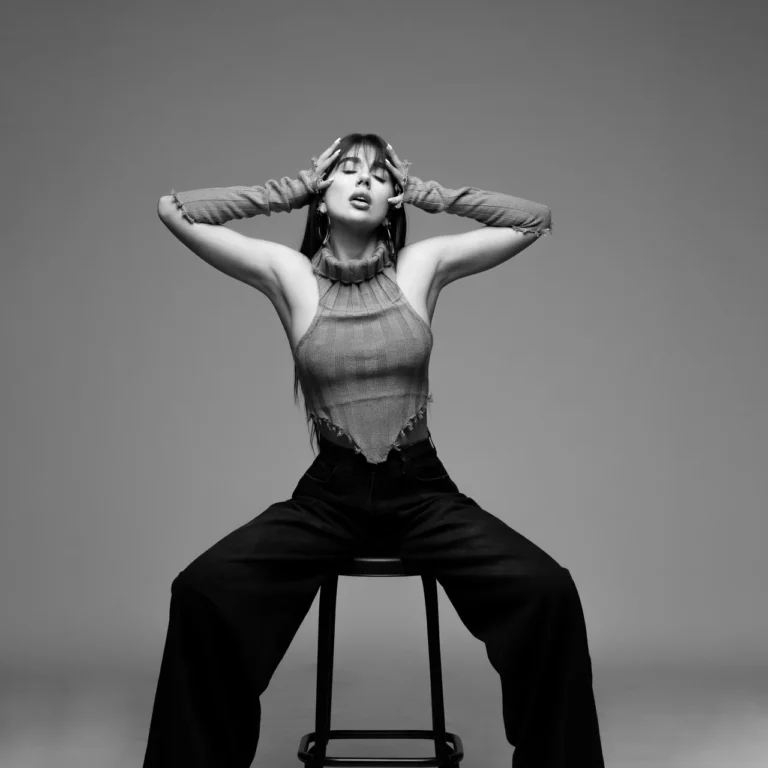 Black-and-white portrait of a woman seated on a stool, hands framing her head, wearing a ribbed sleeveless top and wide-leg pants.