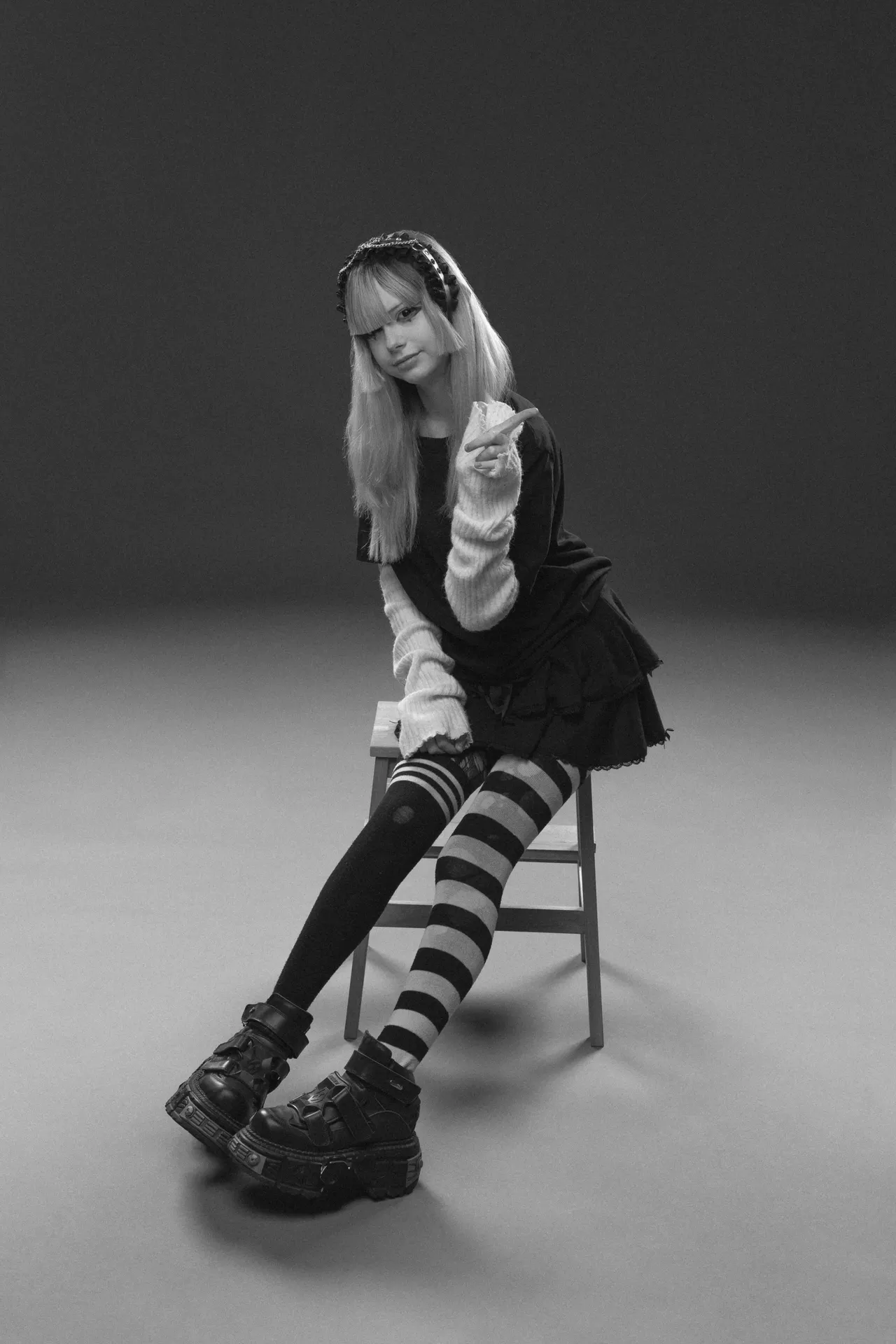 Black-and-white photo of a young woman with long blond hair sitting on a wooden stool, wearing a dark dress, striped tights, and chunky boots, looking at the camera.