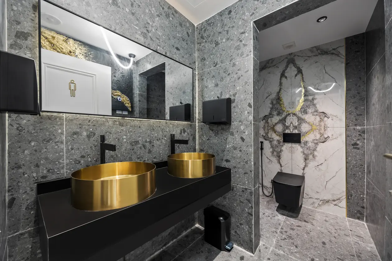 Luxurious bathroom with two gold vessel sinks on a black counter, gray speckled stone walls, and a marble-walled toilet alcove.