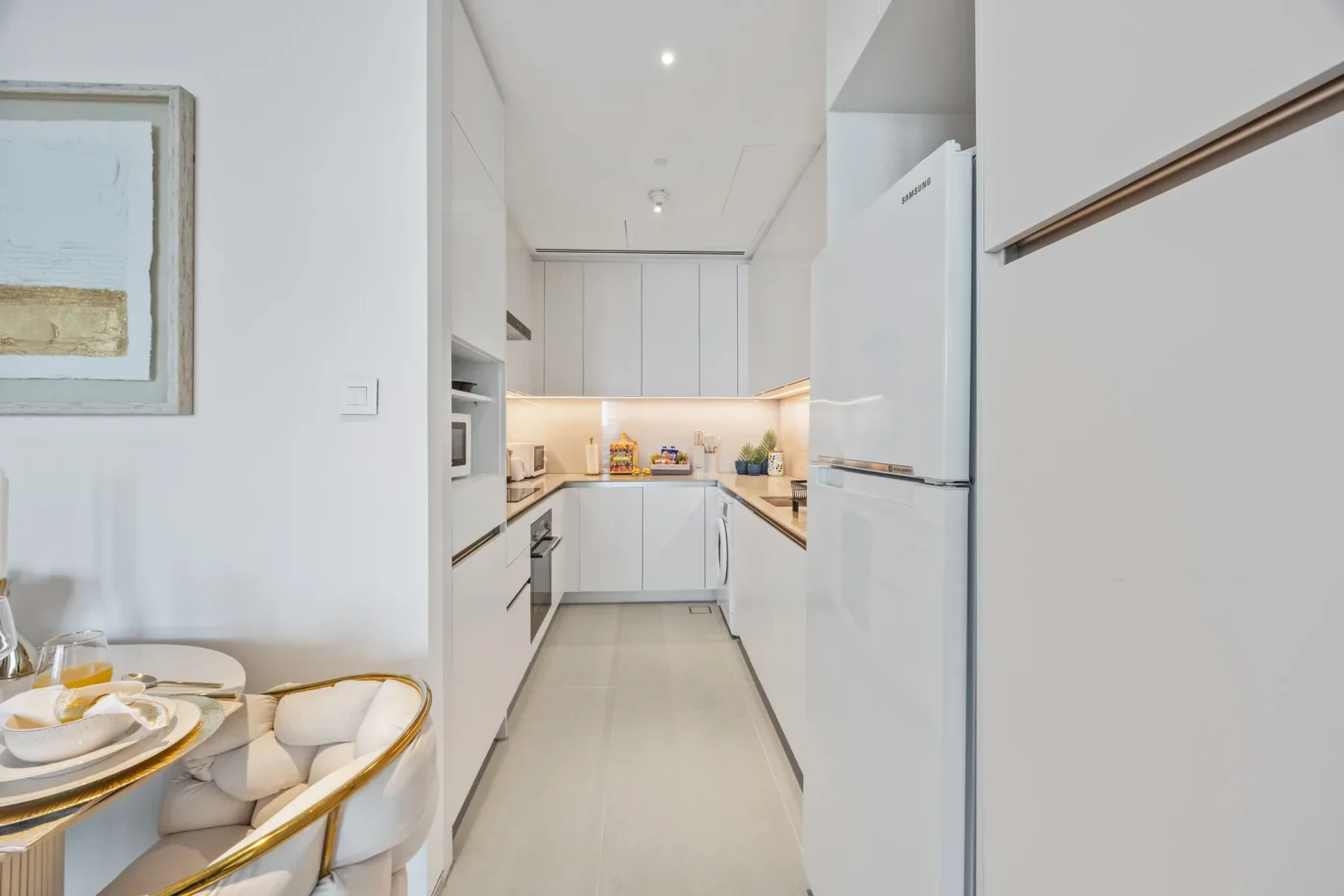 Bright white galley kitchen with built-in appliances, white cabinets, and a Samsung refrigerator, seen from a doorway into the space.
