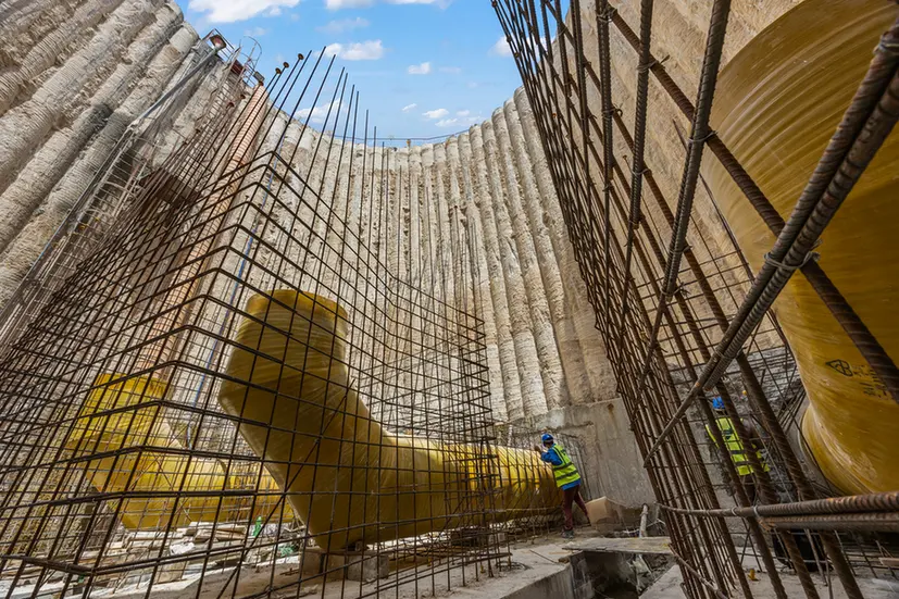 Construction worker in a neon vest inspects a large yellow curved duct inside a concrete circular wall with exposed rebar.