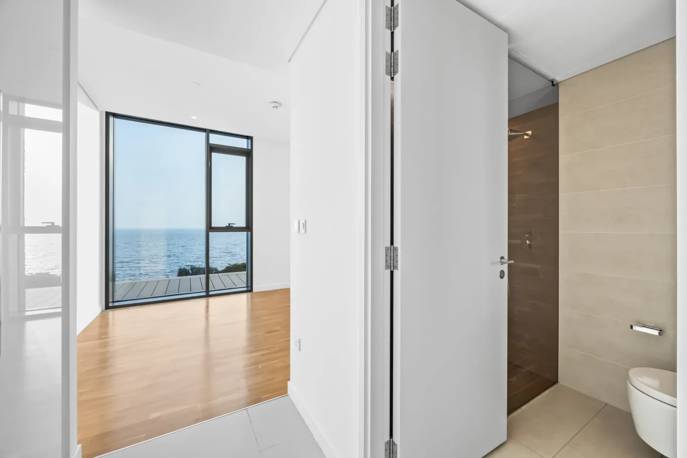 Modern interior opening to a living area with a sea view through large glass doors; bathroom with beige tiles on the right.