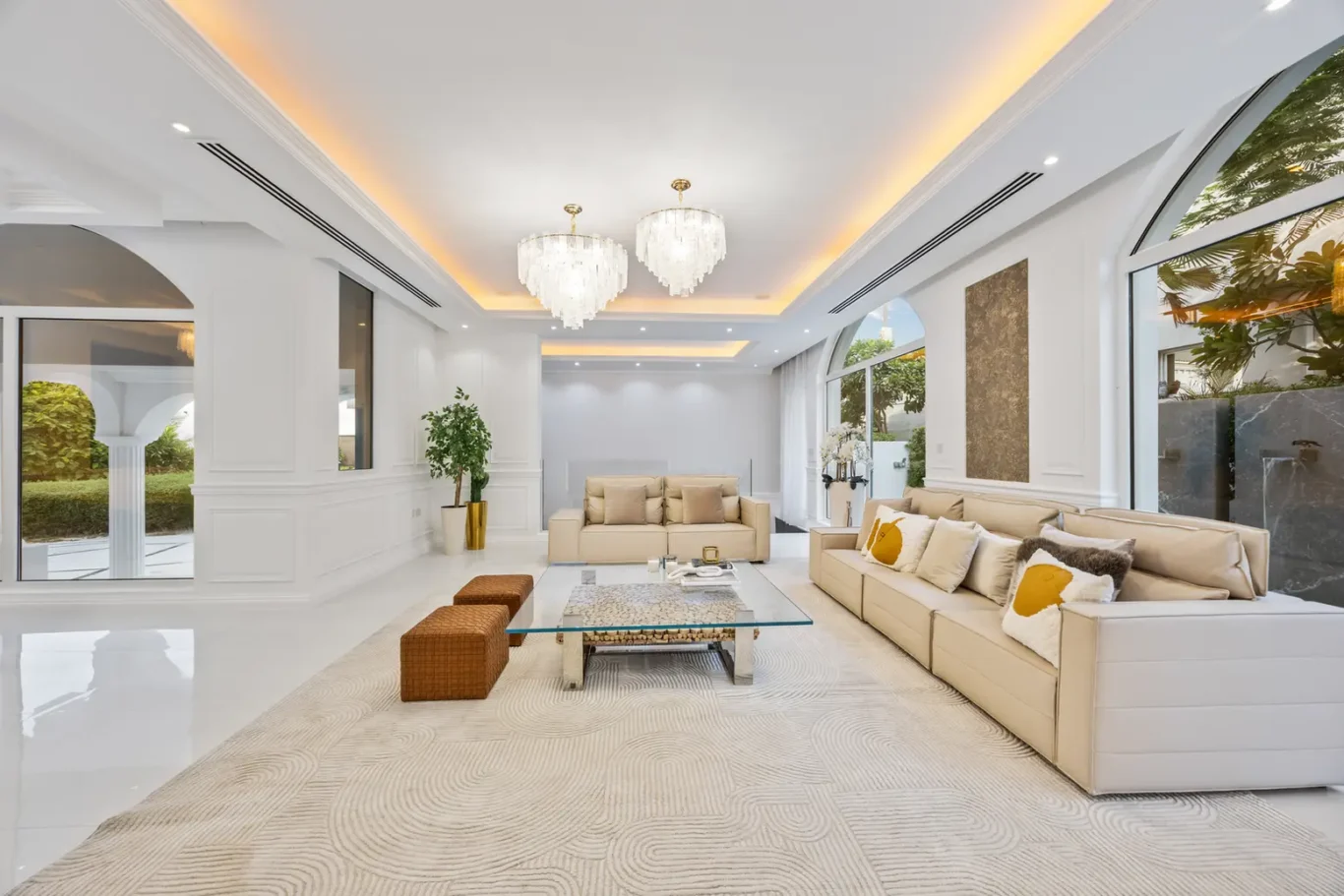 Luxurious bright living room with beige sofas, glass coffee table, and chandeliers, overlooking a garden through large windows.