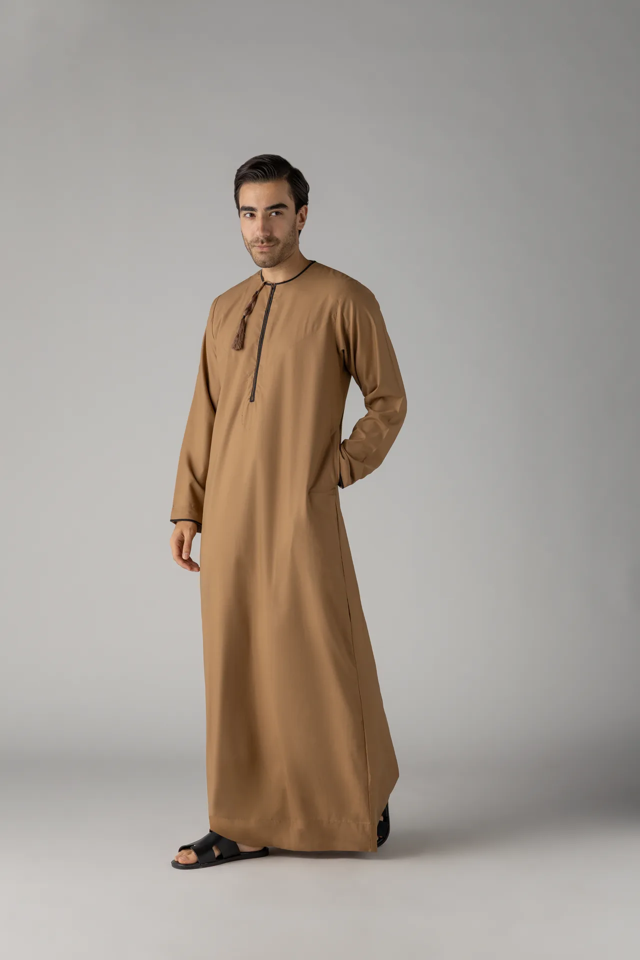 Male model in a tan thobe with a tassel at the neck, standing against a gray studio backdrop, wearing black sandals.