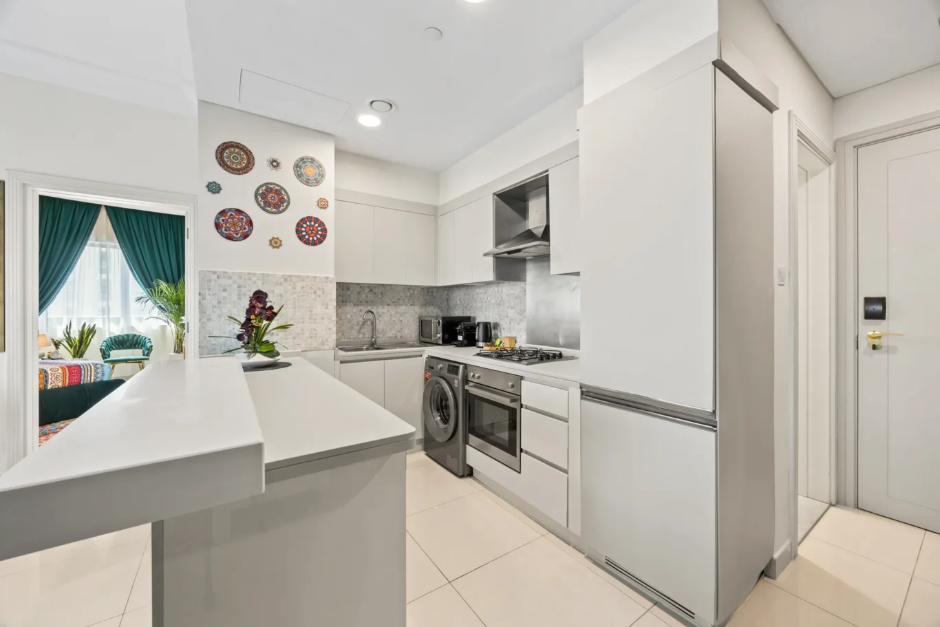 Modern white kitchen with stainless appliances and decorative mosaic backsplash; doorway to a bright seating area with teal curtains outside.