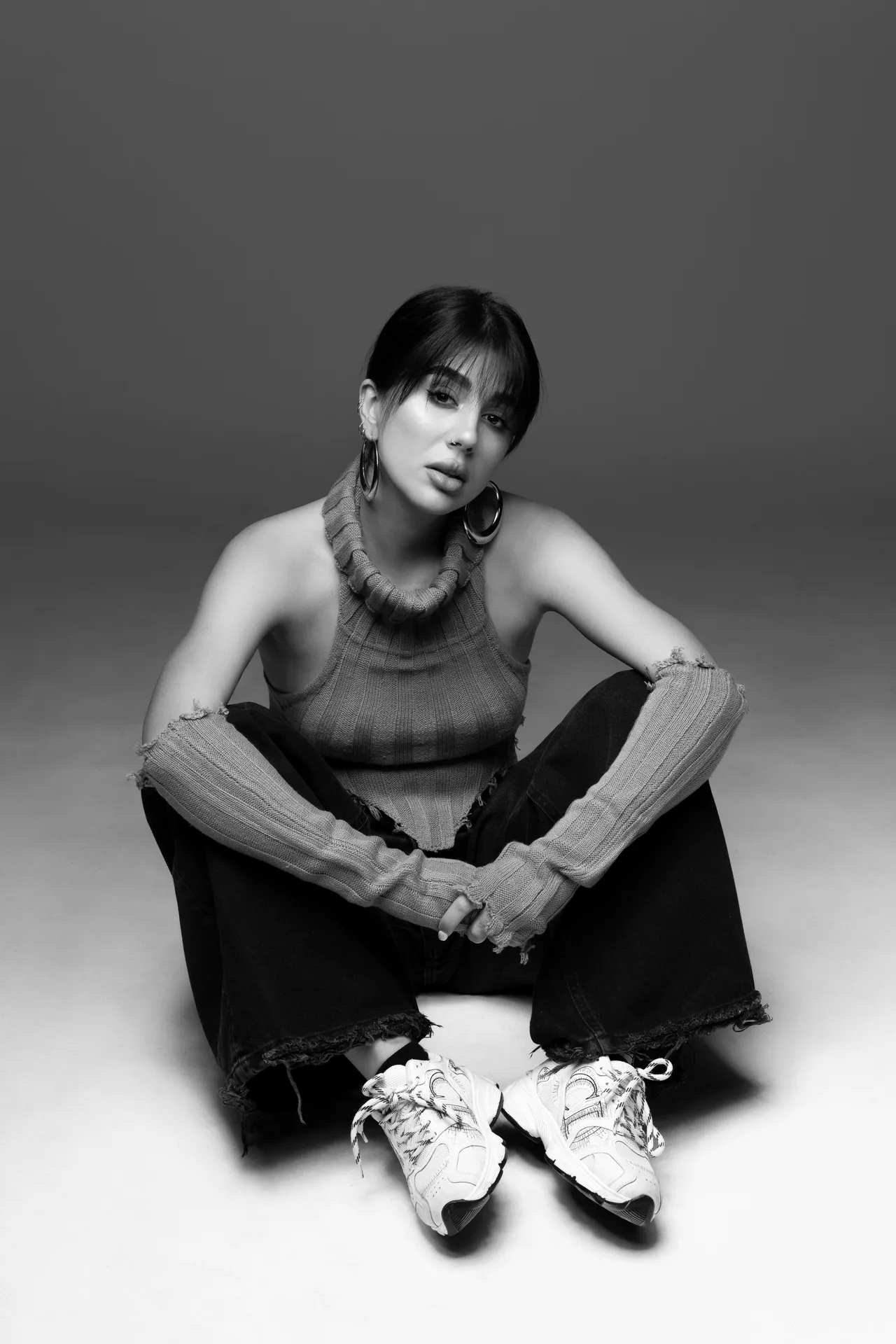 Black-and-white portrait of a young woman seated cross-legged in a studio, wearing a chunky knit sleeveless sweater and large hoop earrings, looking softly at the camera.