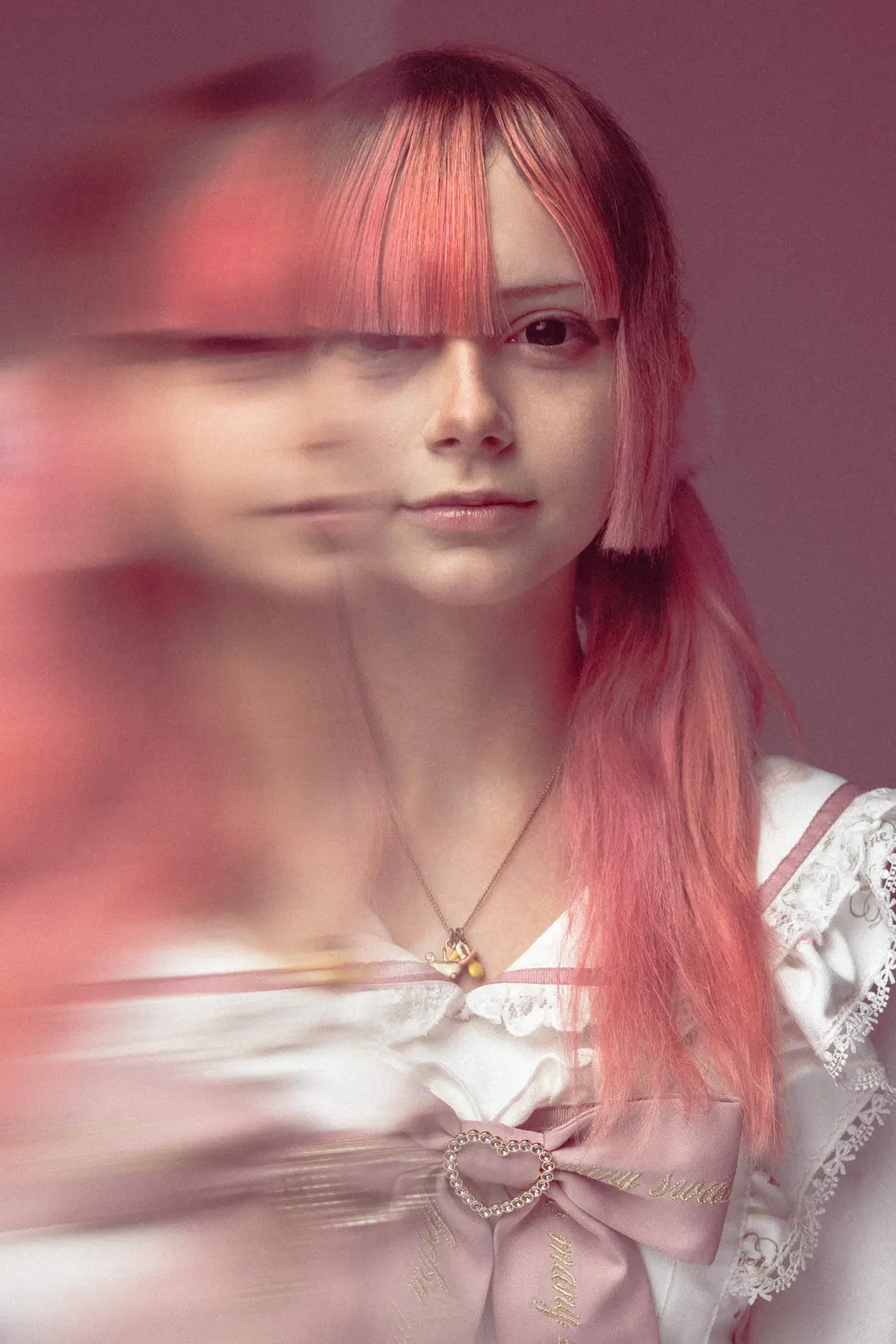 Portrait of a young woman with pink hair and bangs, wearing a white lace top with a pink ribbon and heart-shaped belt buckle, blurred motion on the left side of the frame.
