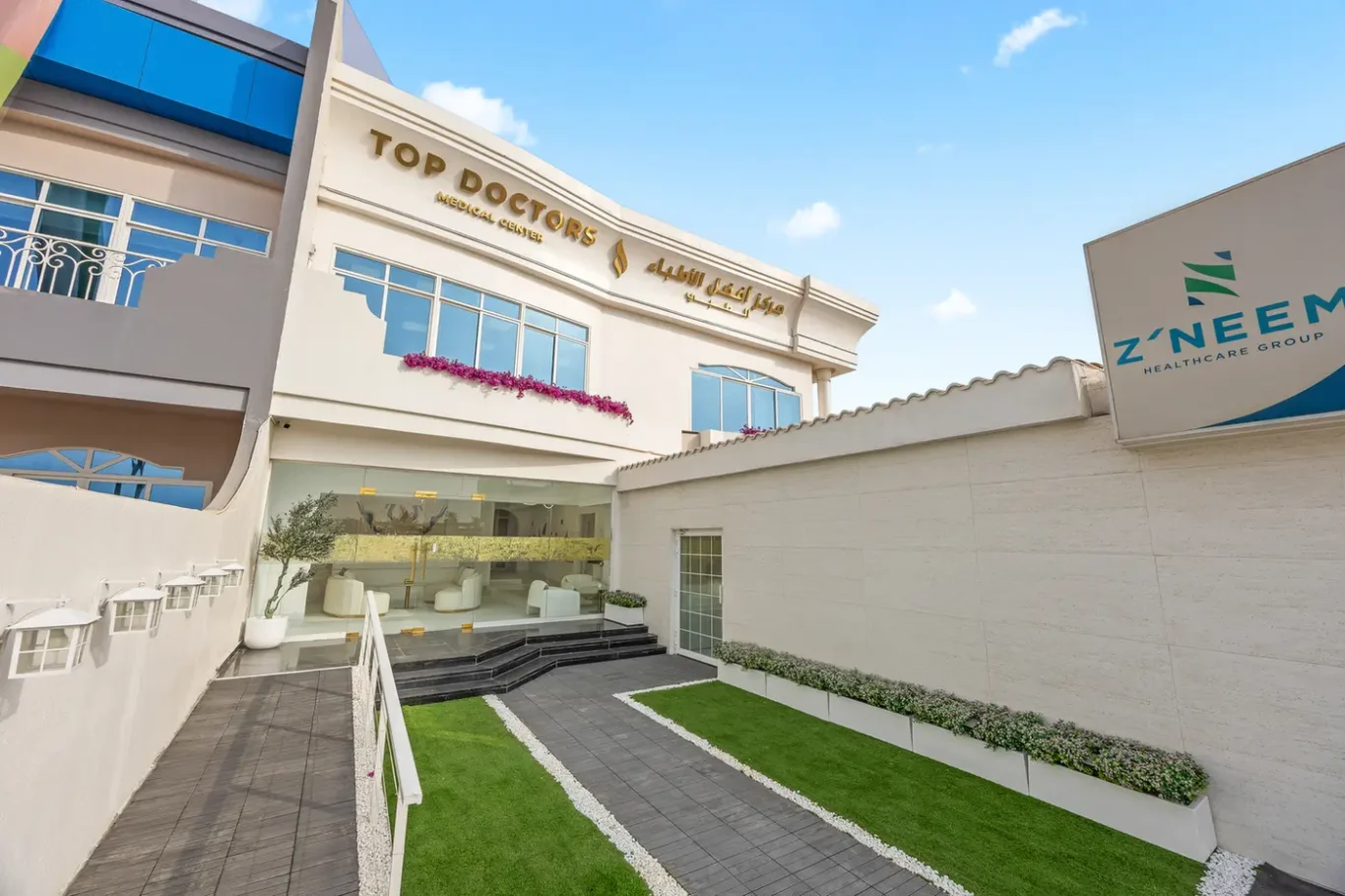 Exterior view of a modern medical center with 'Top Doctors Medical Center' signage and a Z'Neem Healthcare sign on the right side, blue sky above.