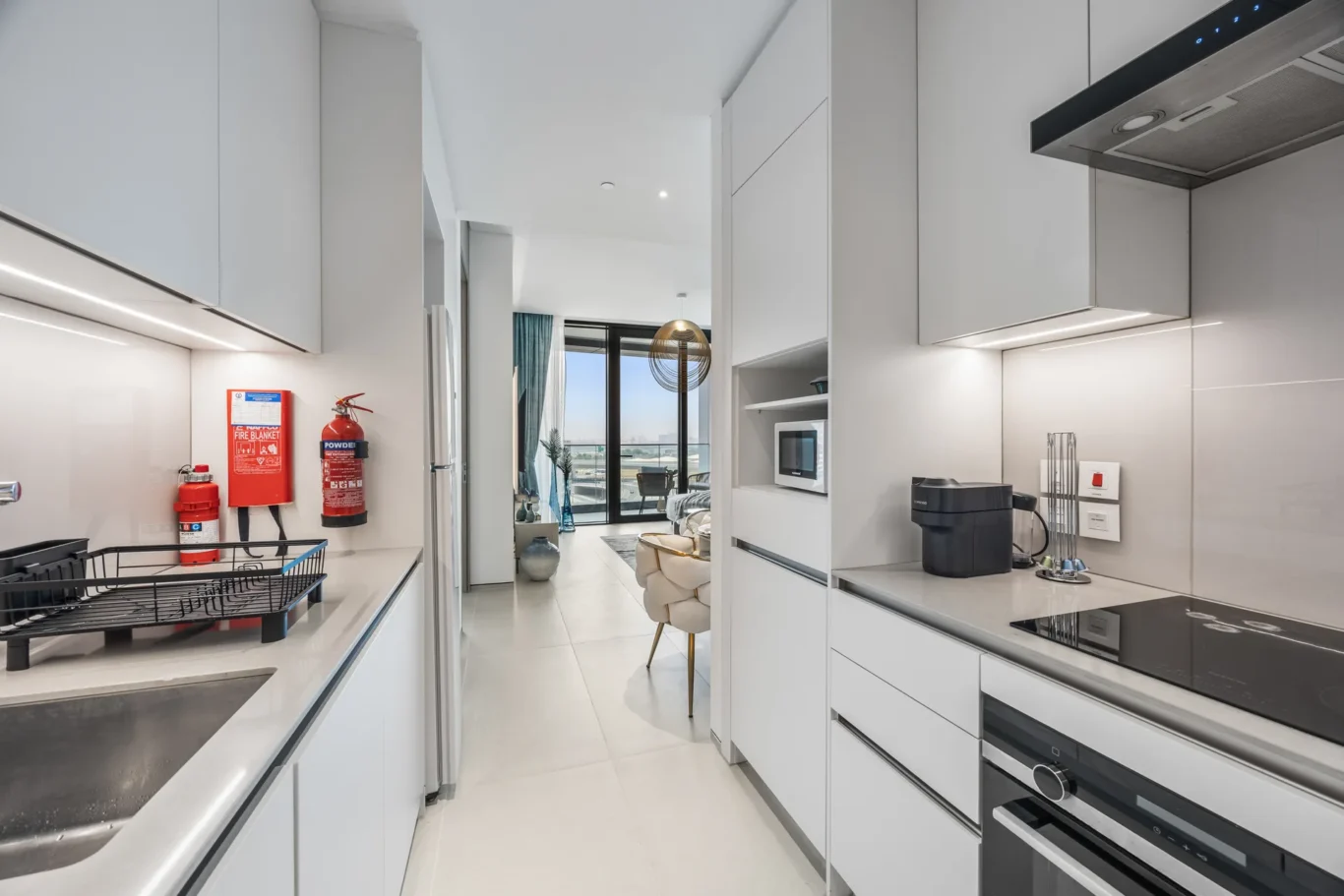 Modern white kitchen with sleek cabinets, stainless countertops, and built-in appliances; a red fire extinguisher hangs on the wall near a dish rack.