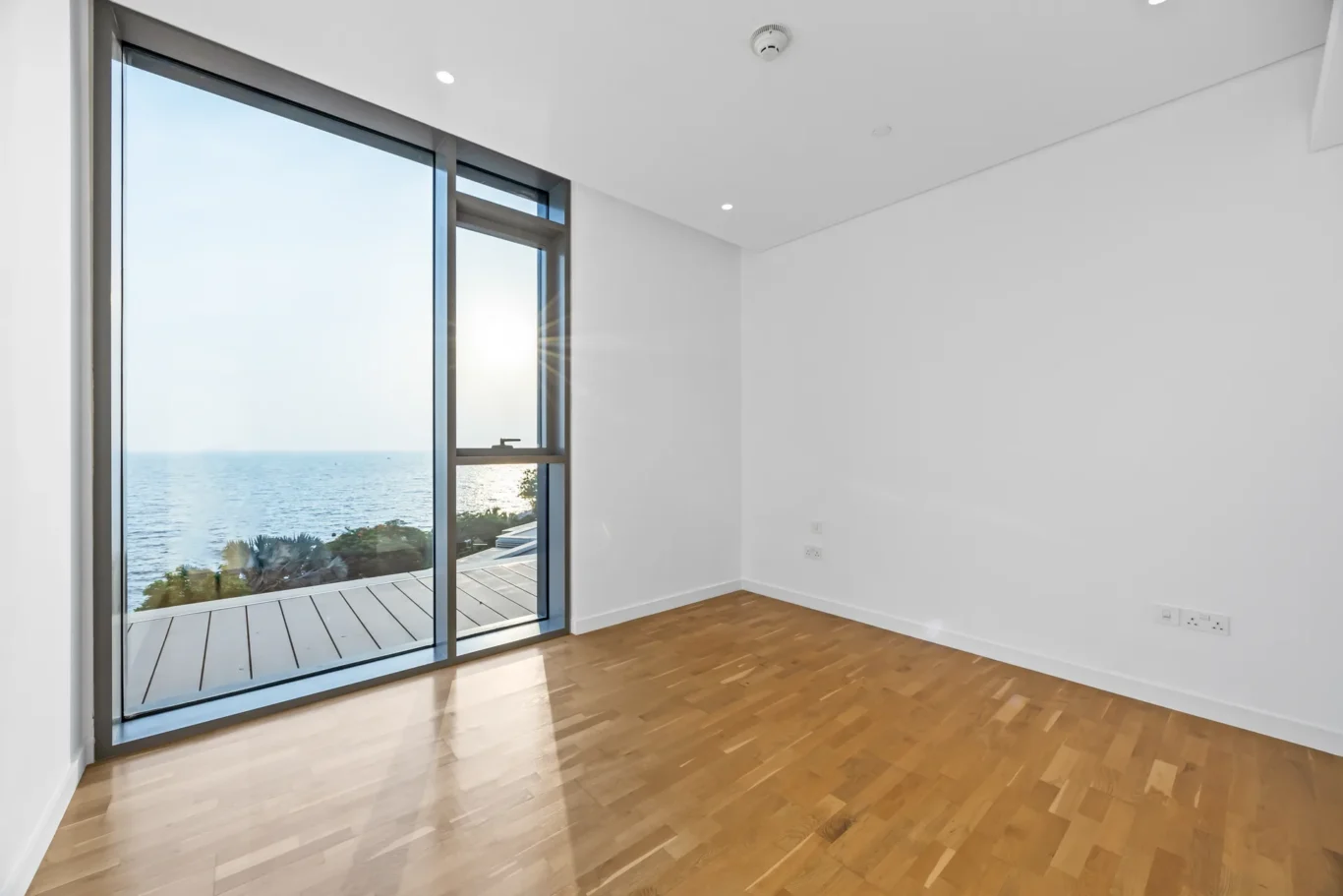 Bright empty room with large sliding glass doors opening to a balcony and ocean view, sun reflecting off the water onto the wood floor.