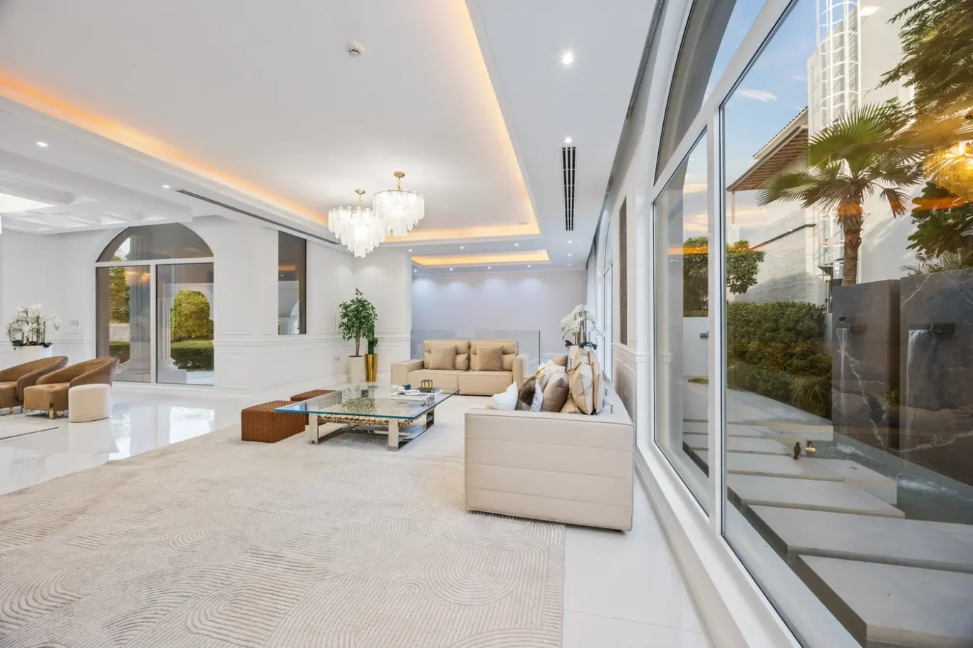 Bright, modern living room with beige sectional sofas, glass coffee table, chandelier, and floor-to-ceiling windows overlooking a garden.
