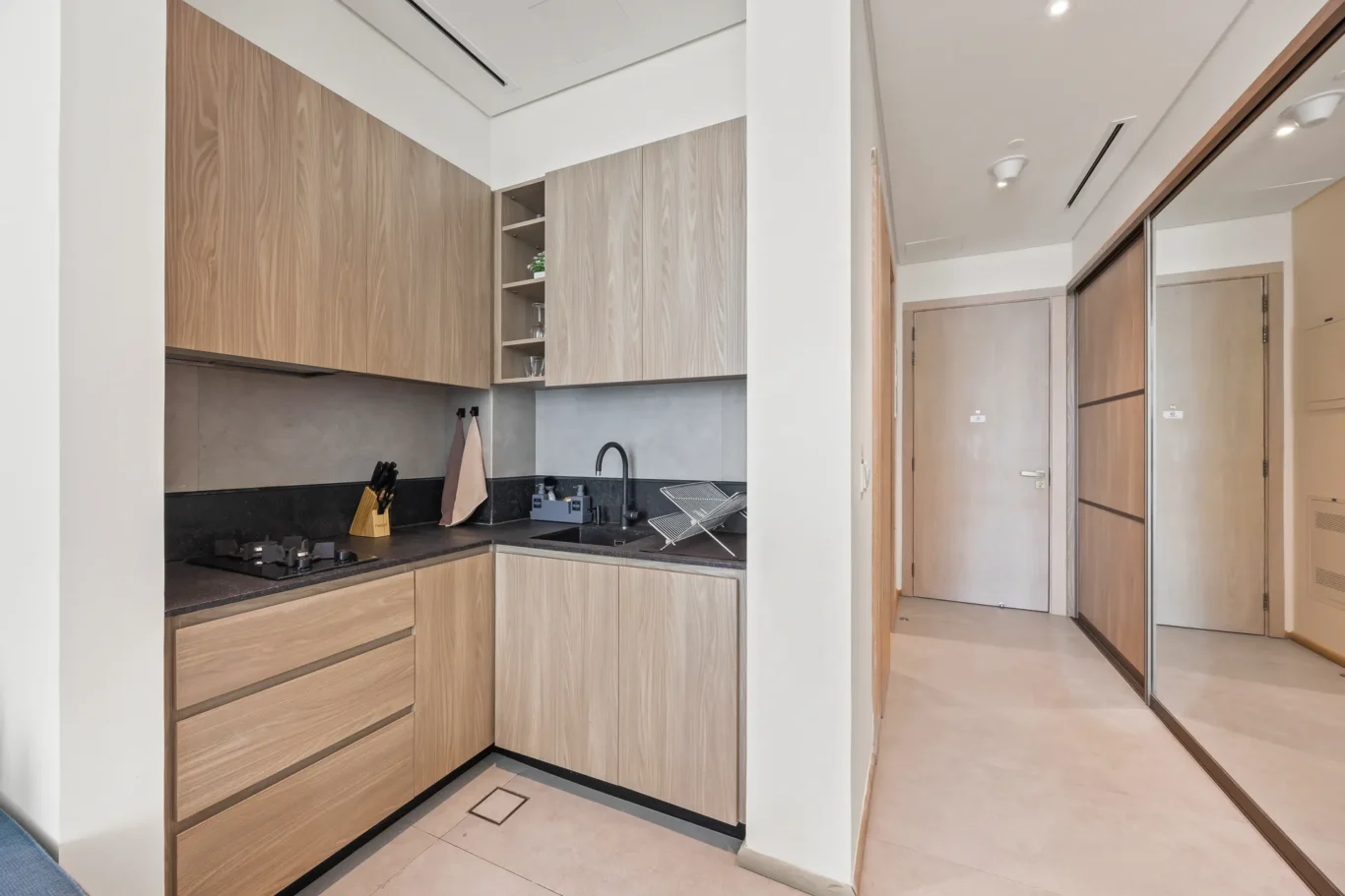 Modern kitchen with light wood cabinetry, black countertop, sink, and gas stove; leads to a hallway with wooden doors.