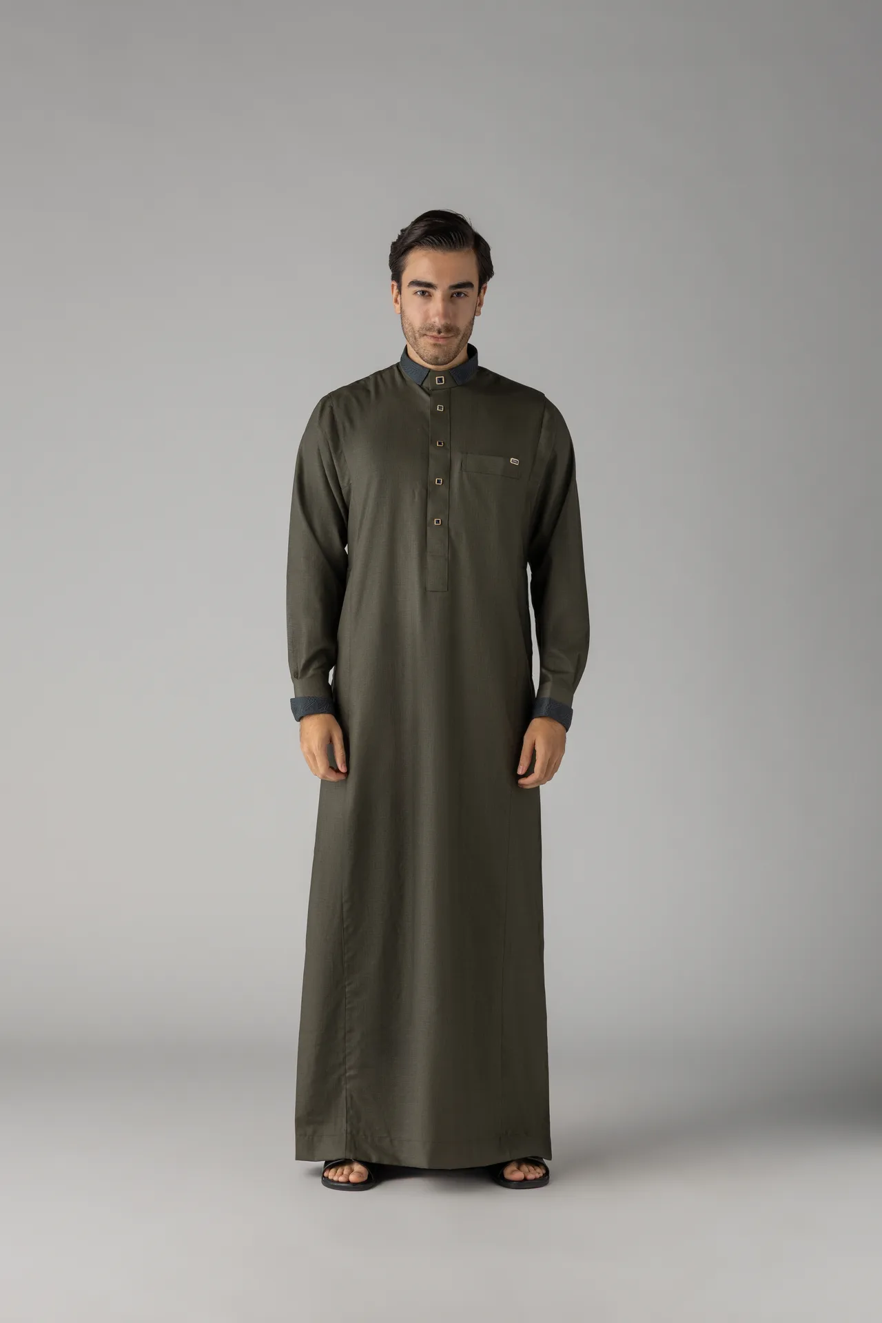 Full-length shot of a man in an olive green thobe standing against a neutral gray backdrop, hands at his sides.
