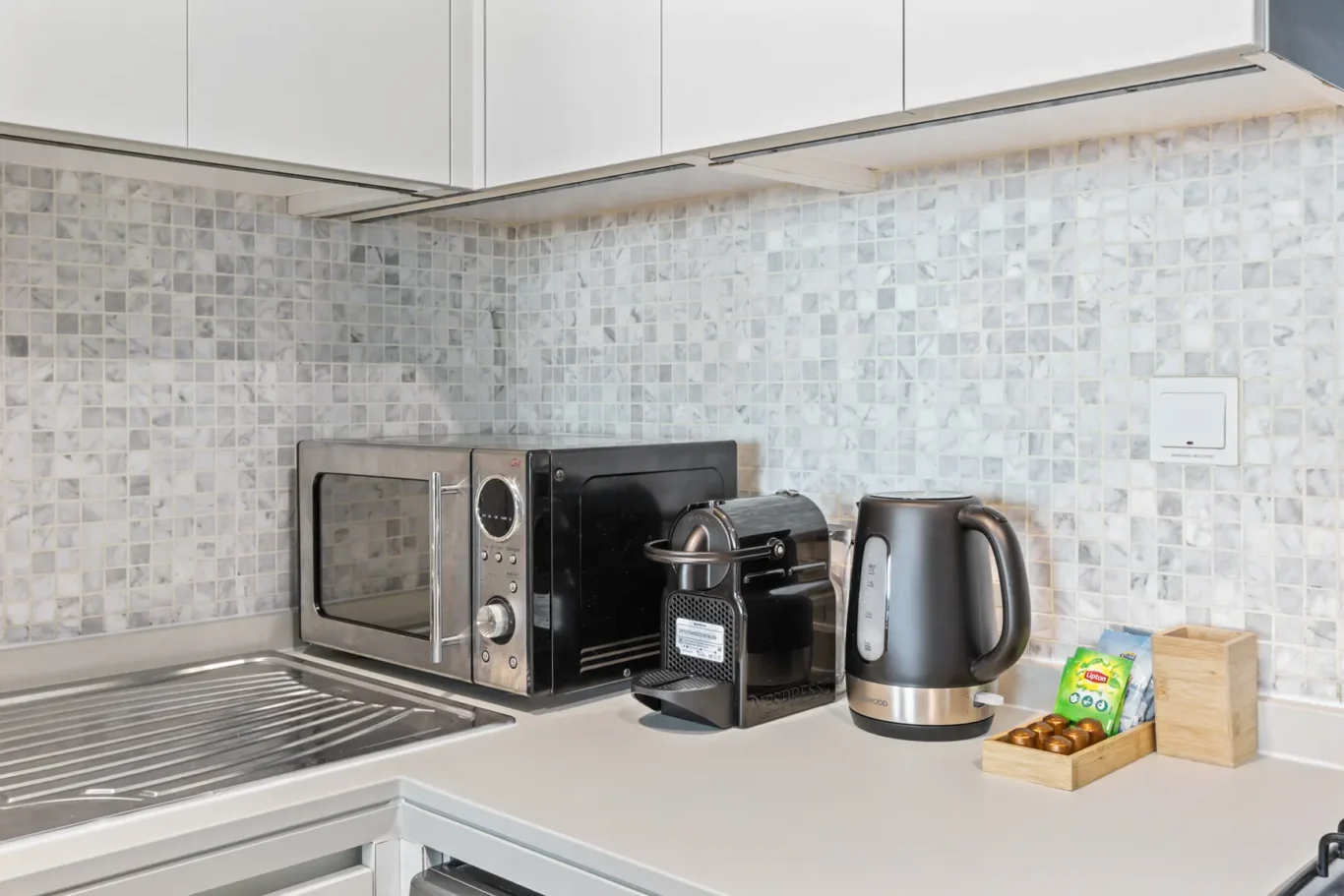 Kitchen counter with microwave, espresso machine, coffee maker, and electric kettle against a tiled backsplash.