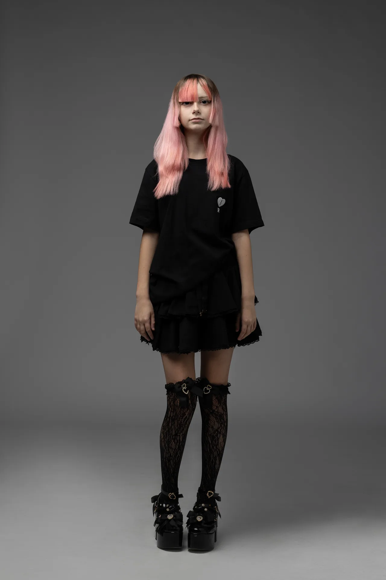 Person standing in a black gothic outfit with pink hair, against a gray studio backdrop, facing the camera.