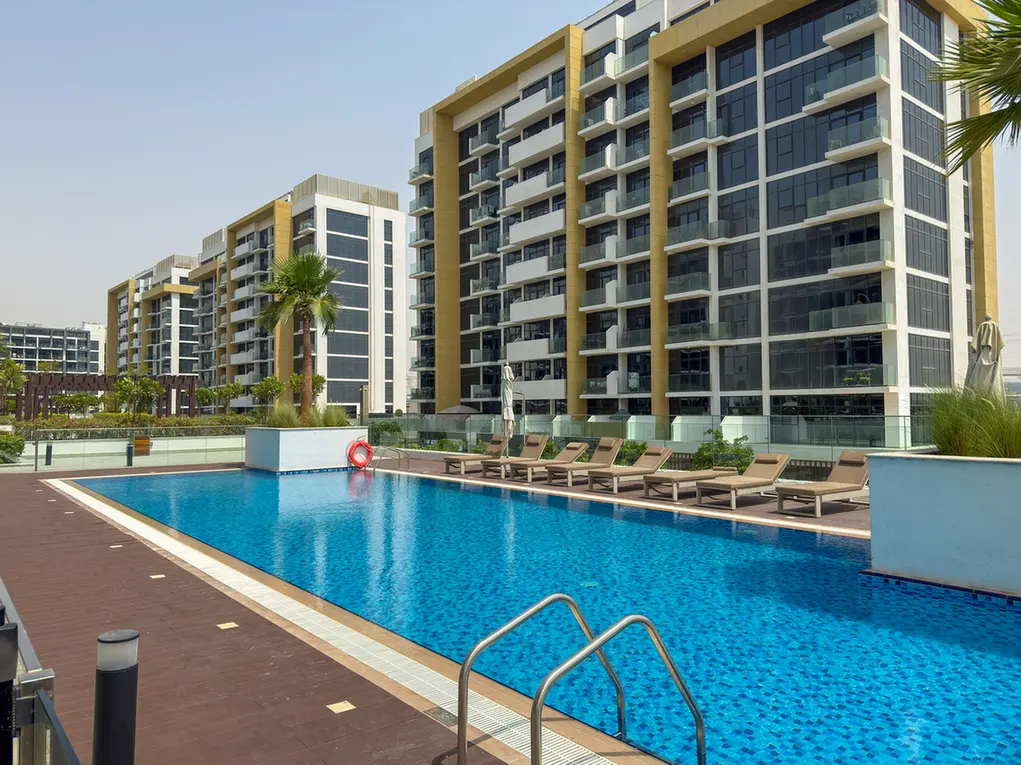 Outdoor pool at a modern residential complex, with lounge chairs, glass railing, and tall glass-front buildings in the background.