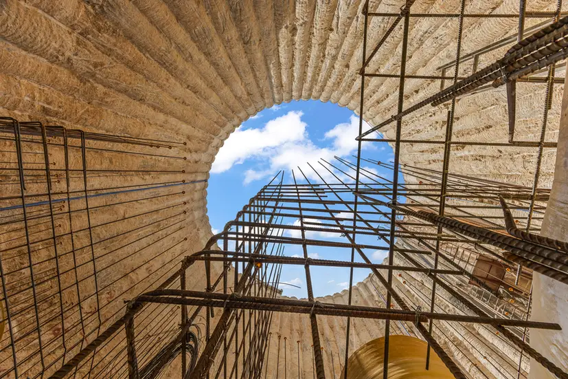 Looking up through a circular brick well under construction, with metal scaffolding reaching toward a blue sky and white clouds.